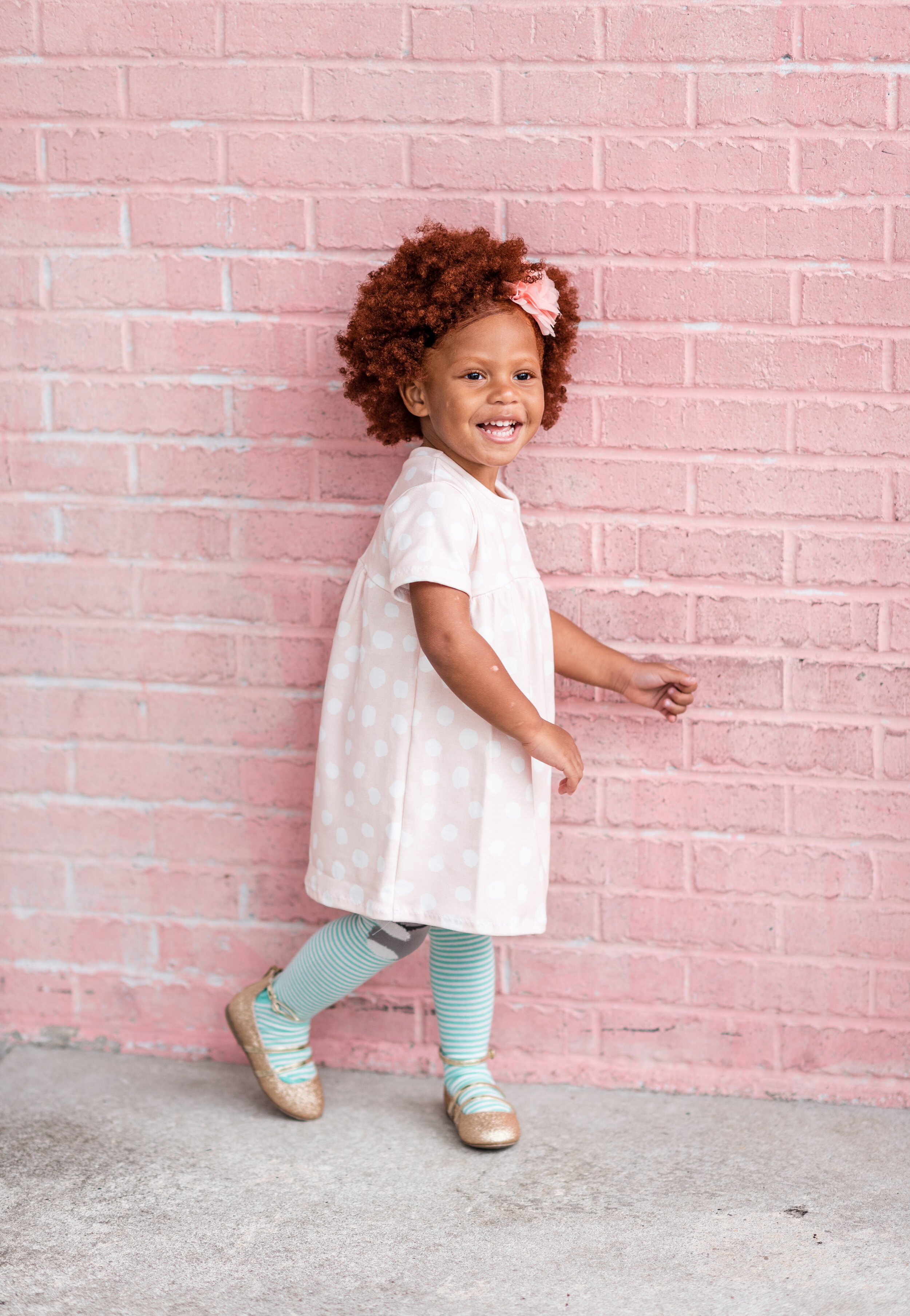A young girl with curly red hair, wearing a pink polka dot dress, striped teal and white tights, and gold shoes, smiling and walking beside a pink brick wall.