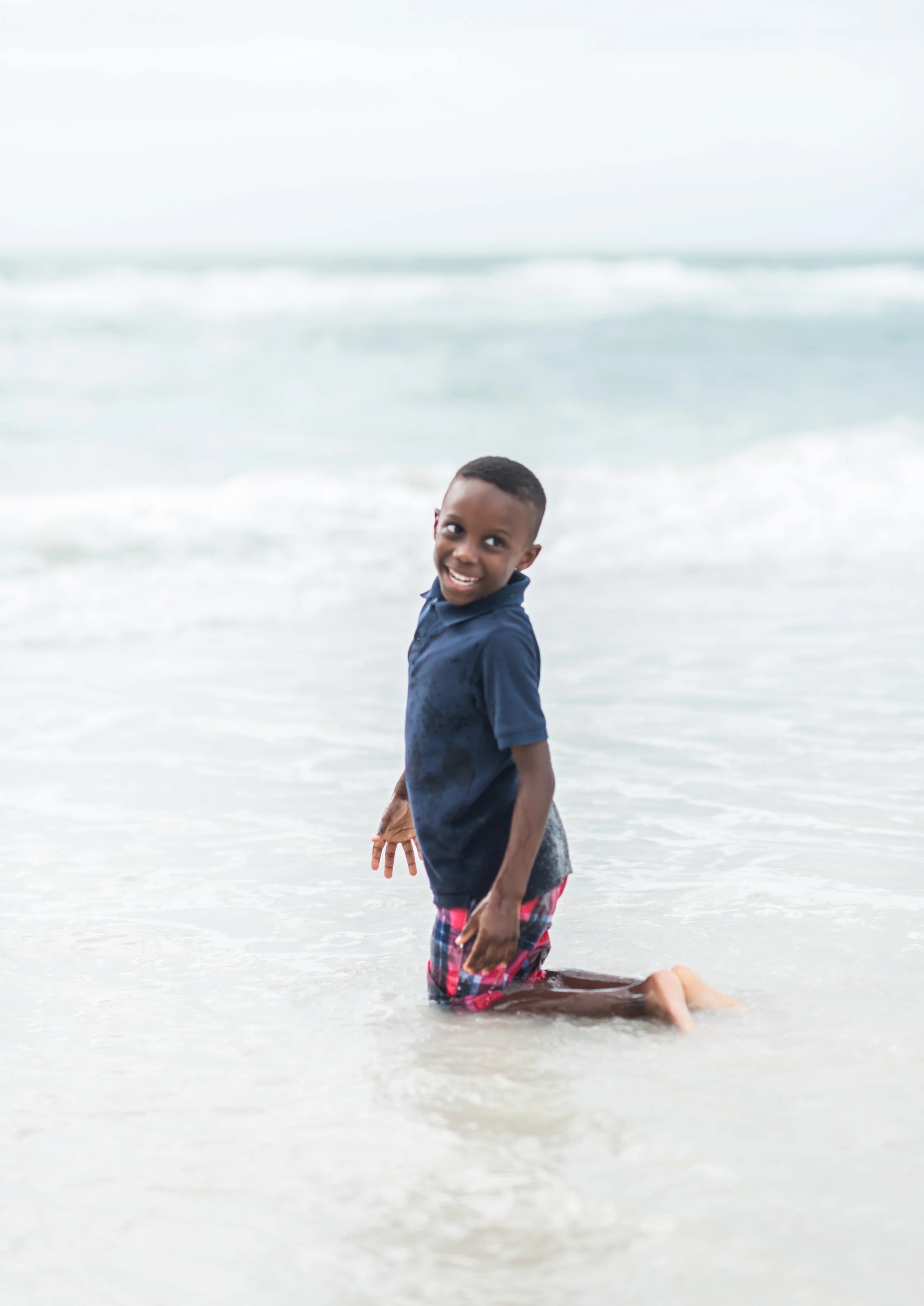 A young boy smiling and playing in shallow ocean water at the beach.