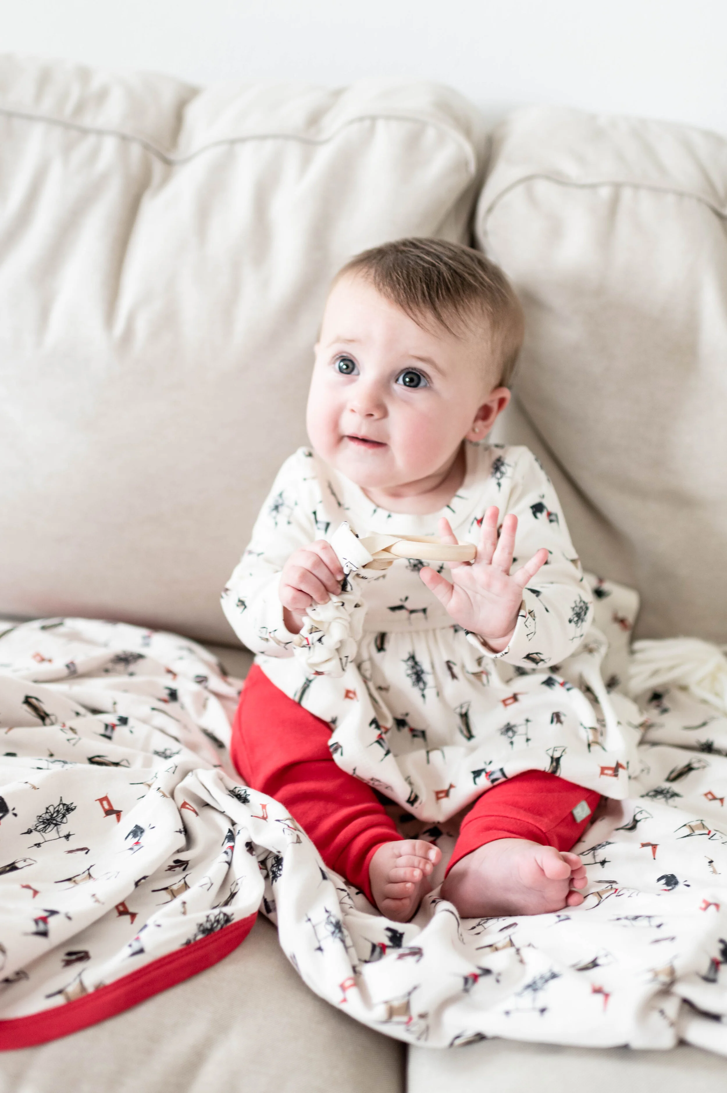 A young child with short brown hair and blue eyes sitting on a beige couch, wearing a white dress with black and red patterns and red pants, holding a wooden spoon and raising one hand.