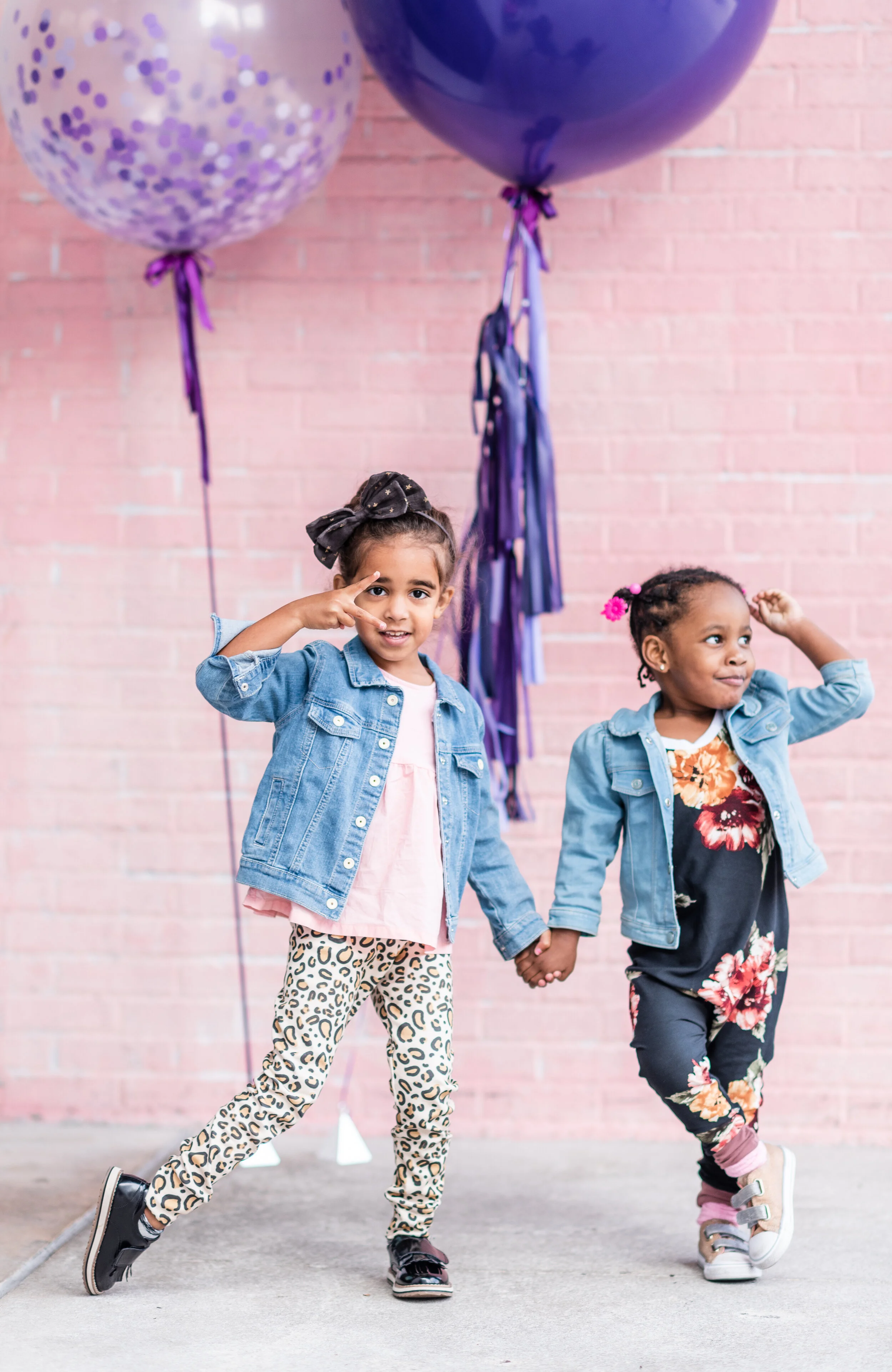 Two young girls holding hands at a celebration in front of pink brick wall and purple balloons, one girl making a peace sign and the other scratching her head.