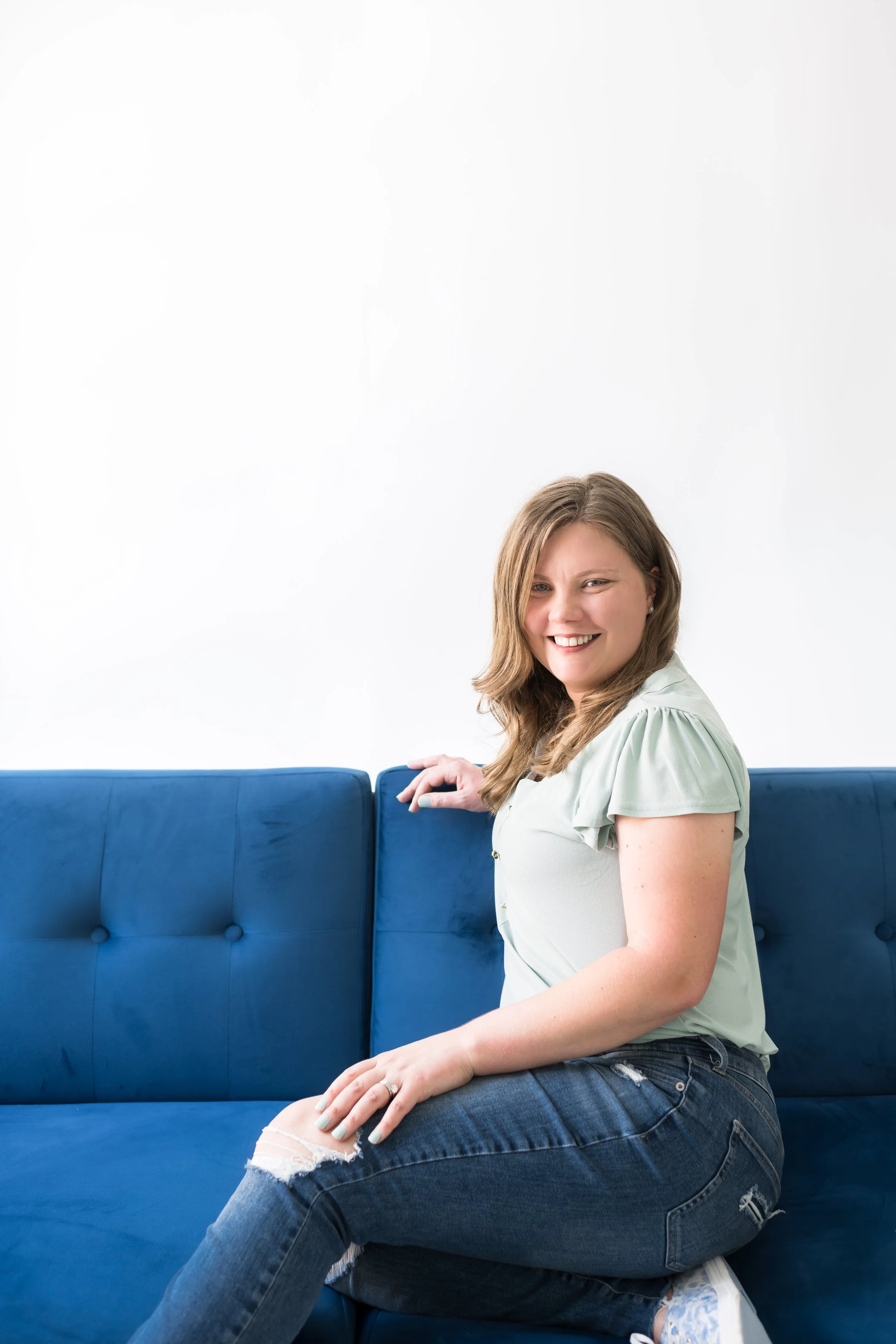 Young woman with light brown hair smiling, sitting on a blue velvet sofa with a white wall background.