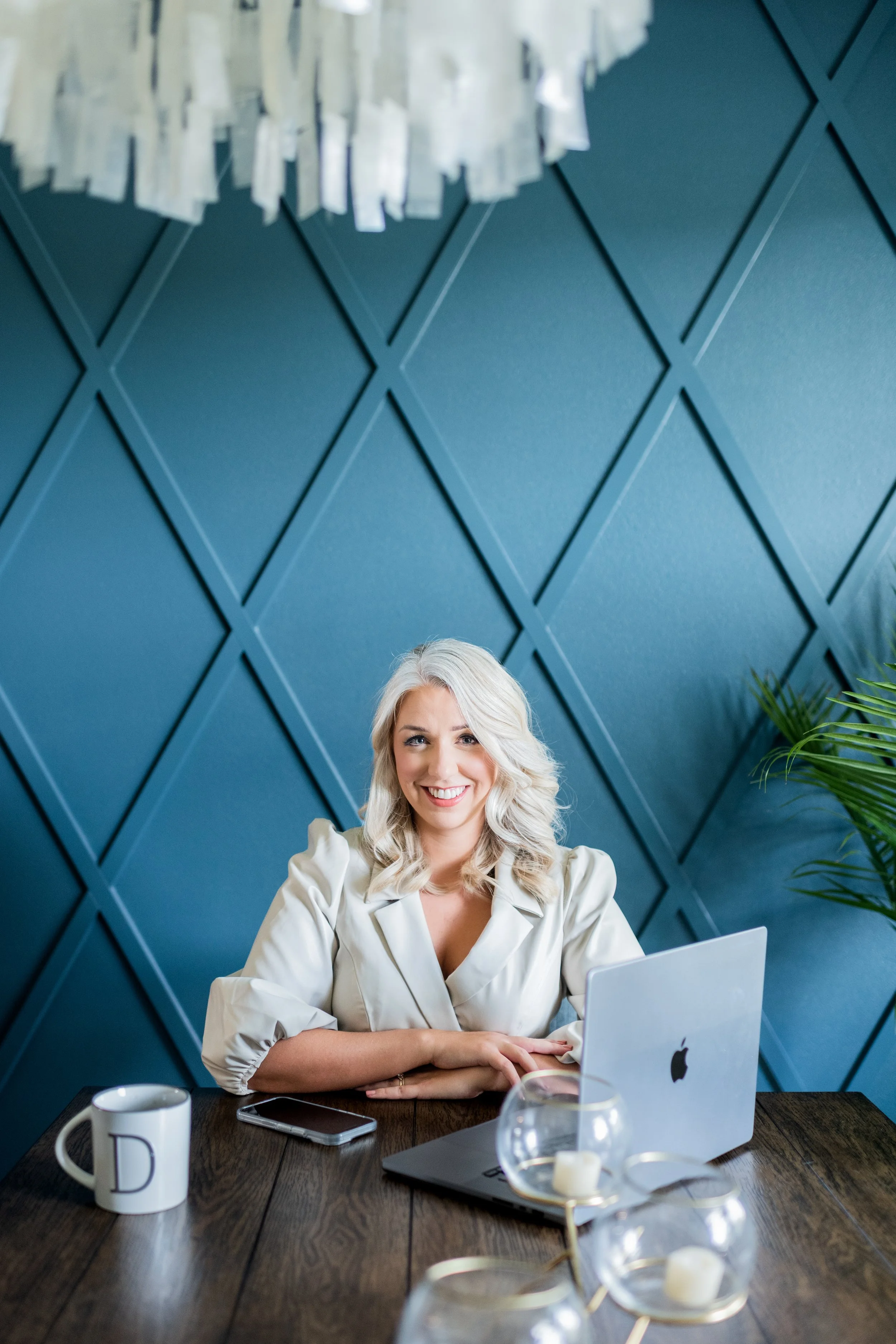 A woman with blonde hair sits at a wooden table with a silver laptop, coffee mug, and smartphone, smiling at the camera in a modern office with a blue geometric wall and green plants.