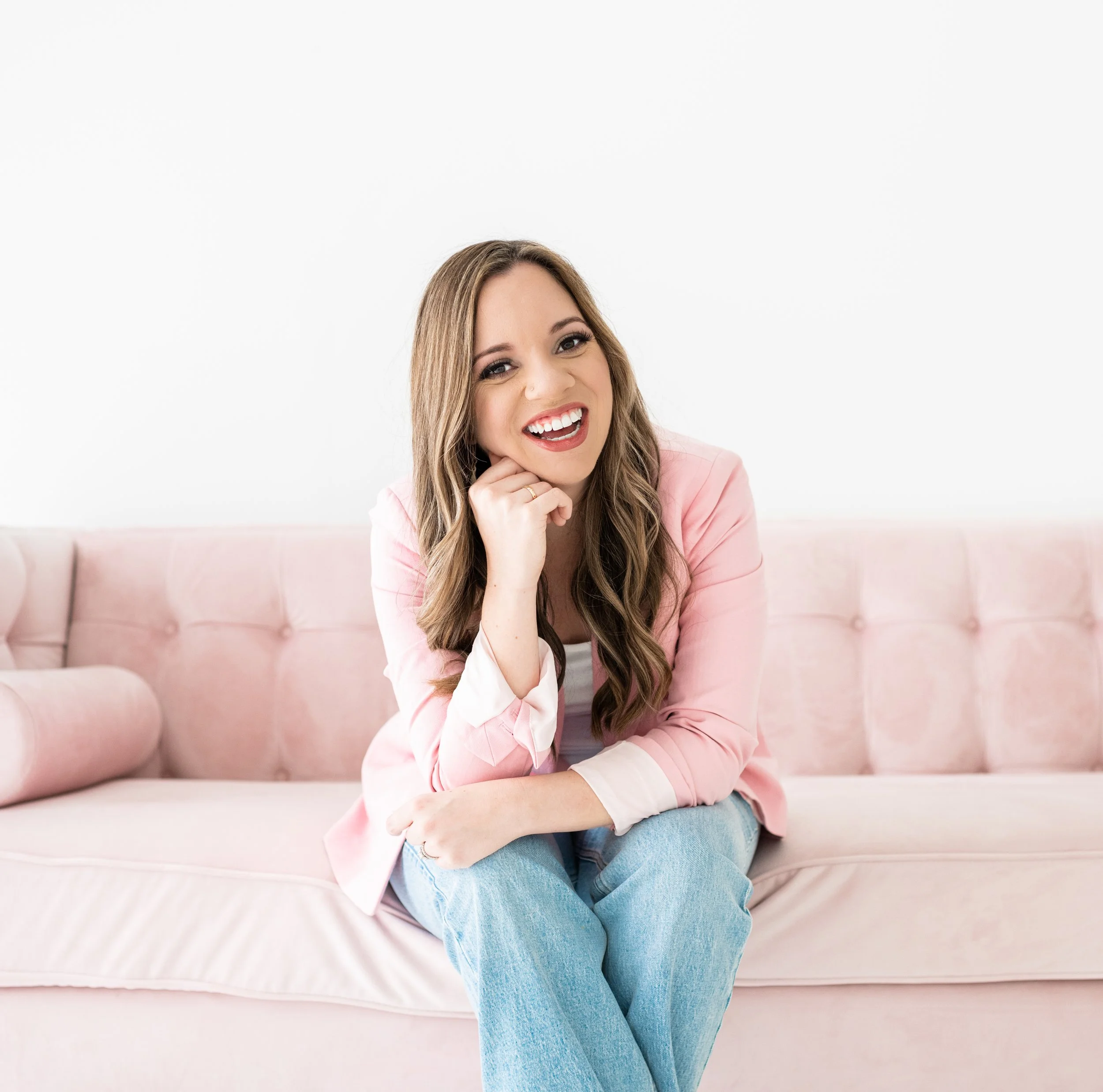 A smiling woman with long wavy hair sitting on a pink tufted sofa, wearing a pink blazer and light jeans, posing with her chin resting on her hand against a plain white background.
