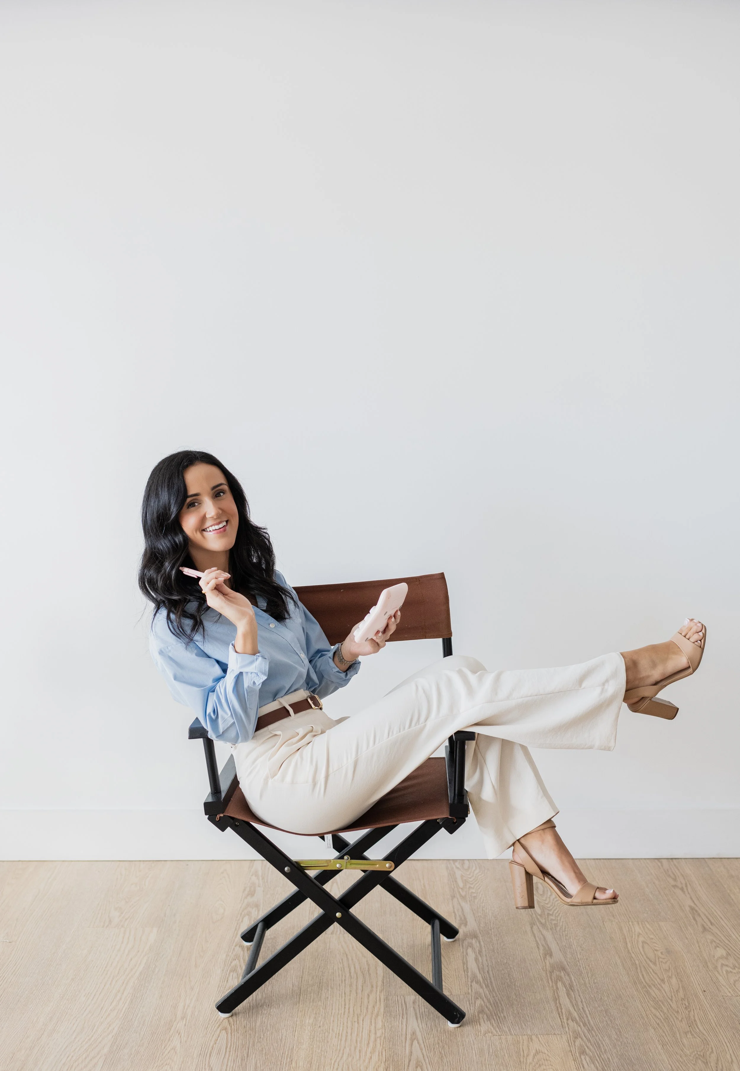 A woman sitting on a foldable chair, smiling, holding a pen and a smartphone, wearing a light blue blouse, cream-colored pants, and tan high heels, against a plain white wall.