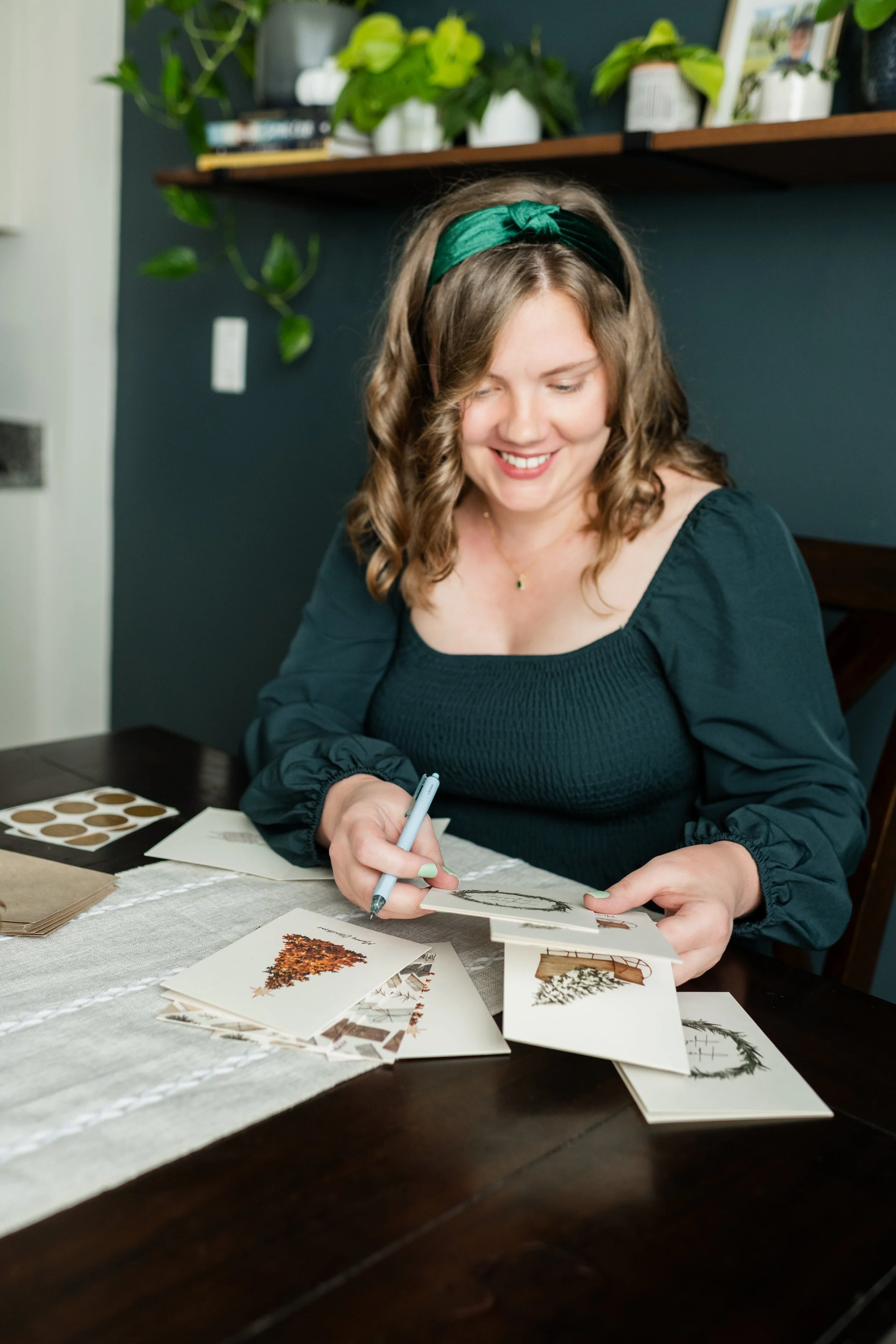 A woman with wavy brown hair, wearing a green headband and a black long-sleeve top, is sitting at a dark wooden table, smiling while she signs holiday cards. The table has several cards with winter and holiday-themed images, and a few sheets of paper