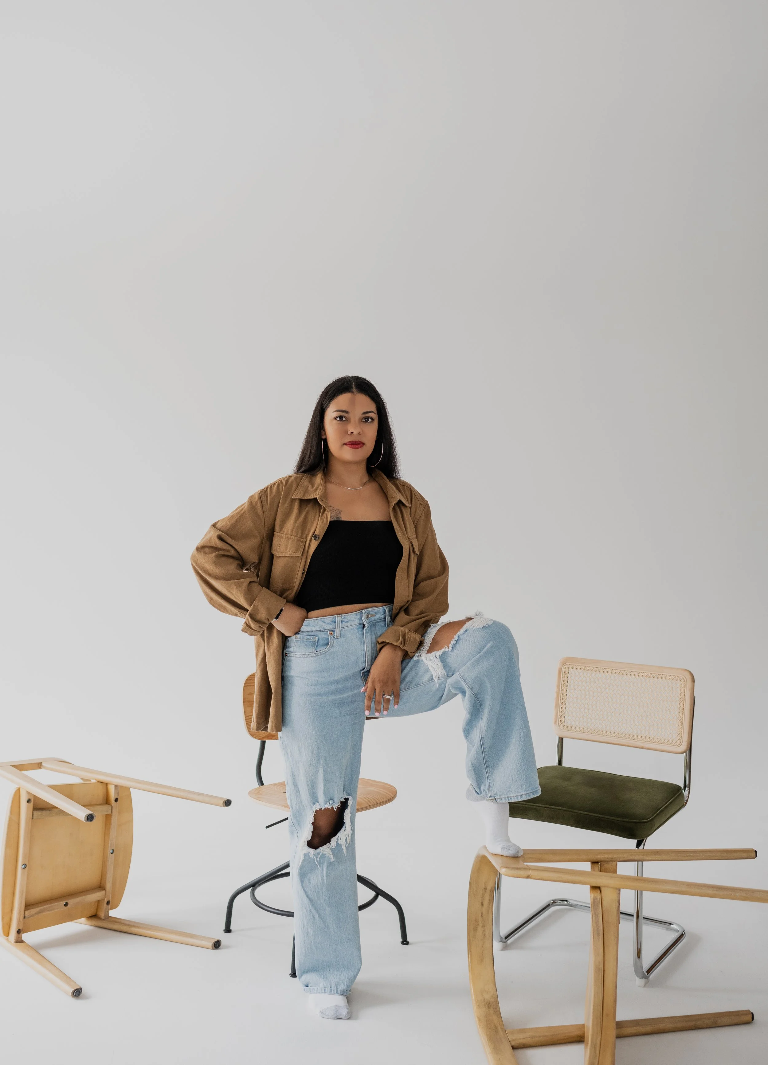 A woman with long dark hair wearing a black crop top, a tan oversized jacket, and ripped light blue jeans, standing with one foot on a chair, in a white studio surrounded by various empty chairs and a small table.