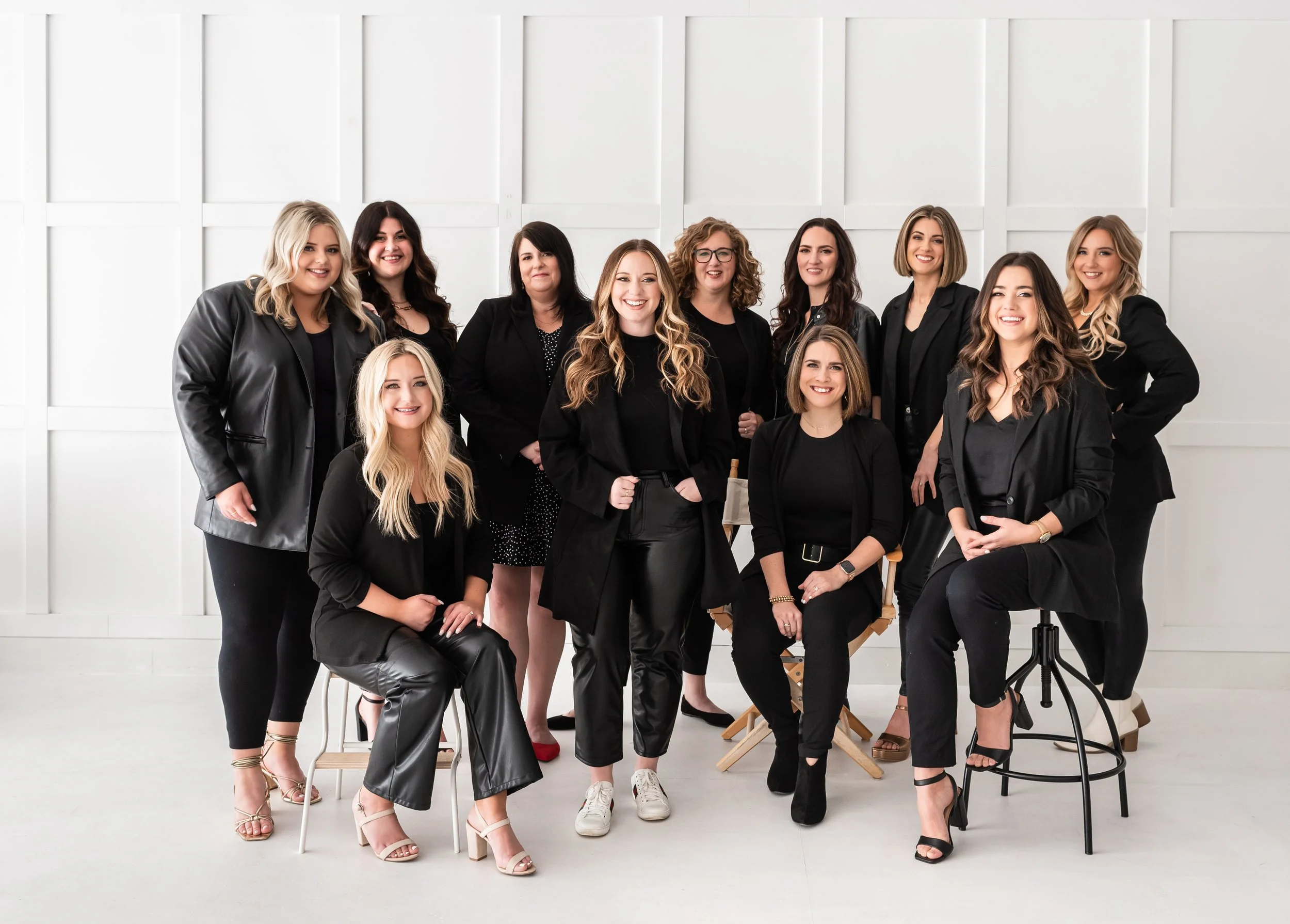 Group of eleven women in black and dark clothing posing together in a bright, white studio space.