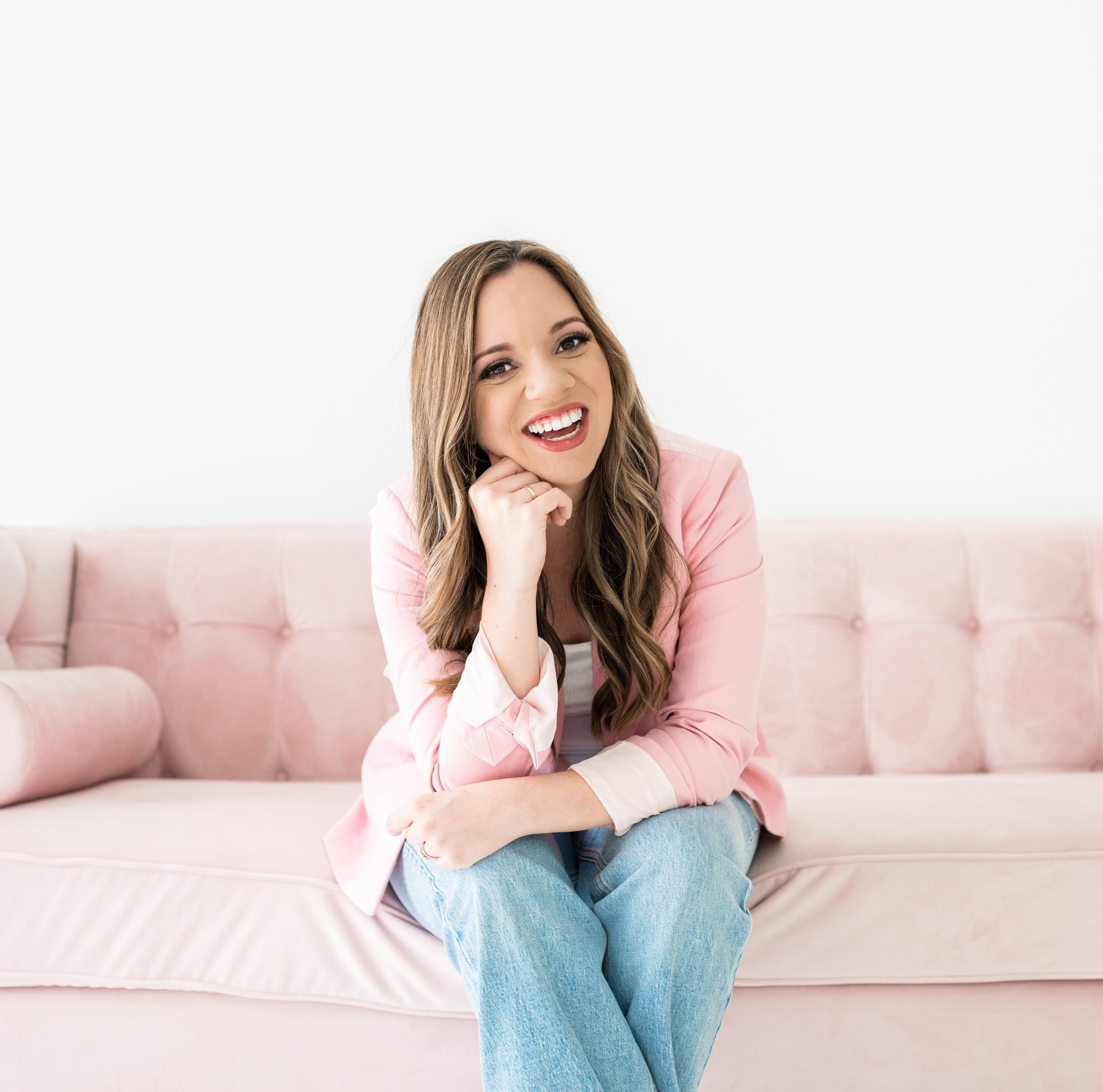 A woman with long wavy hair wearing a pink blazer and light blue jeans sitting on a light pink tufted sofa, smiling at the camera.