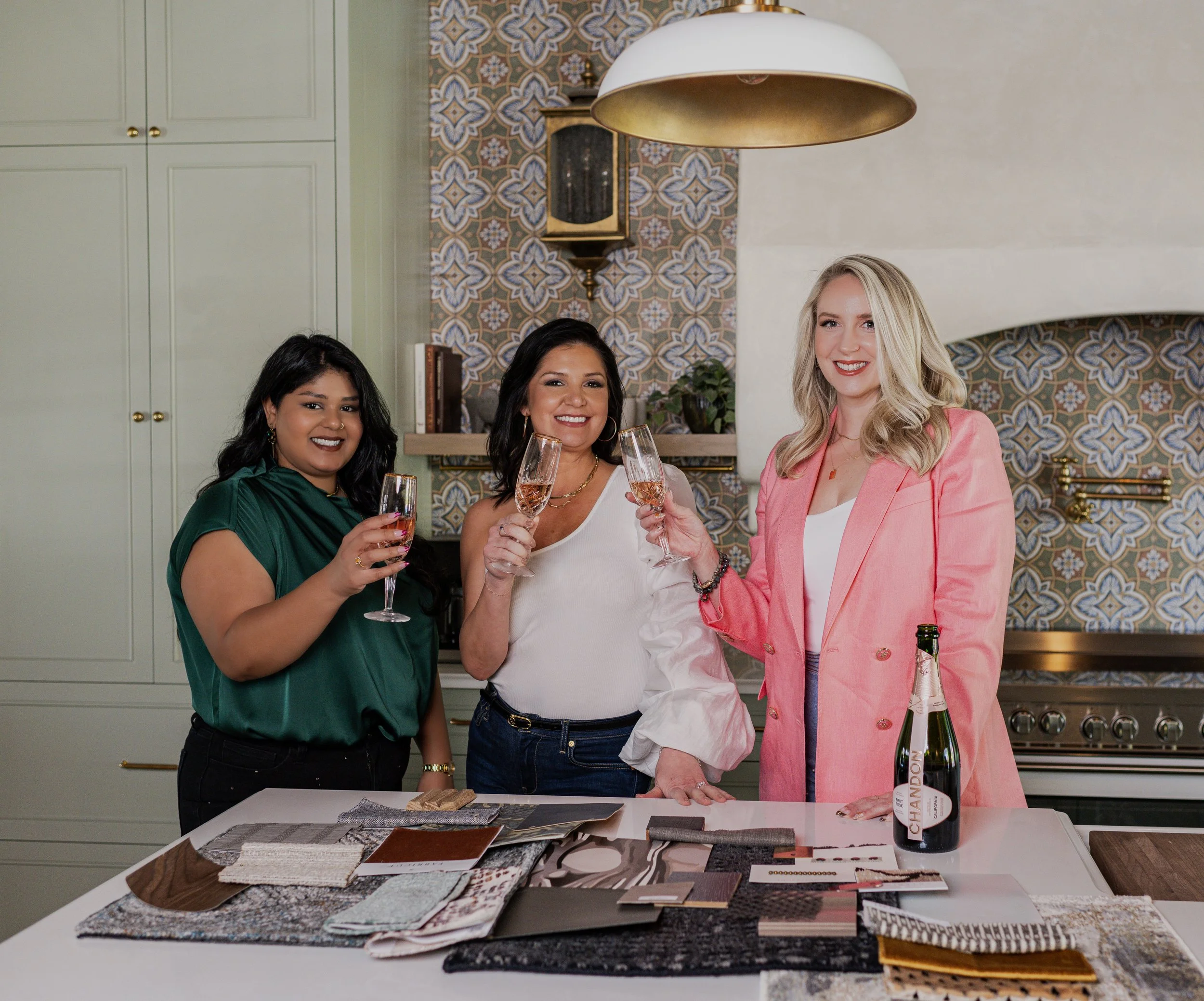 Three women smiling and toasting with glasses of rosé wine in a kitchen, with fabric and design samples spread on the table in front of them.