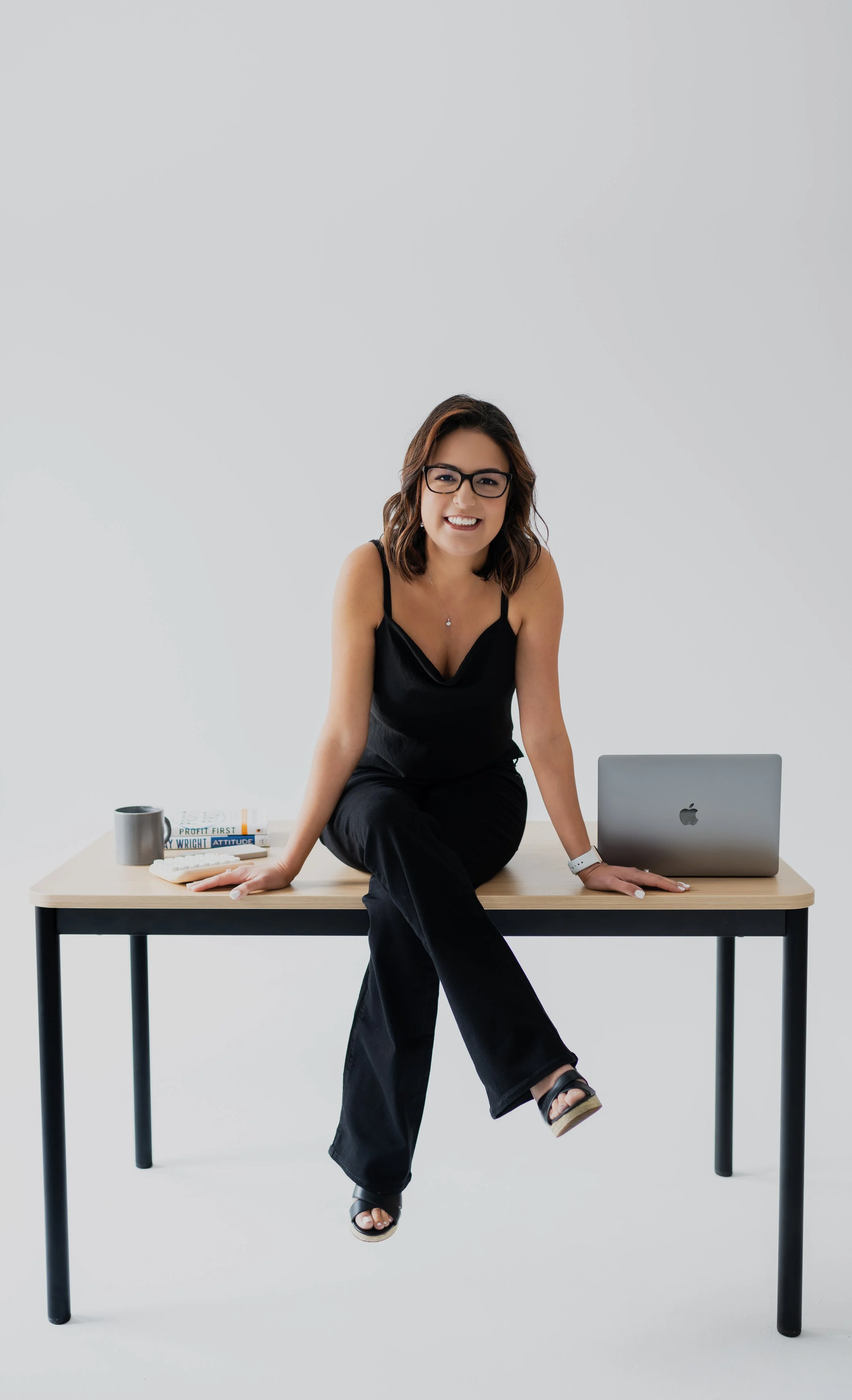 Woman in black clothing sitting on a desk with a laptop, books, and a coffee mug, smiling at the camera.