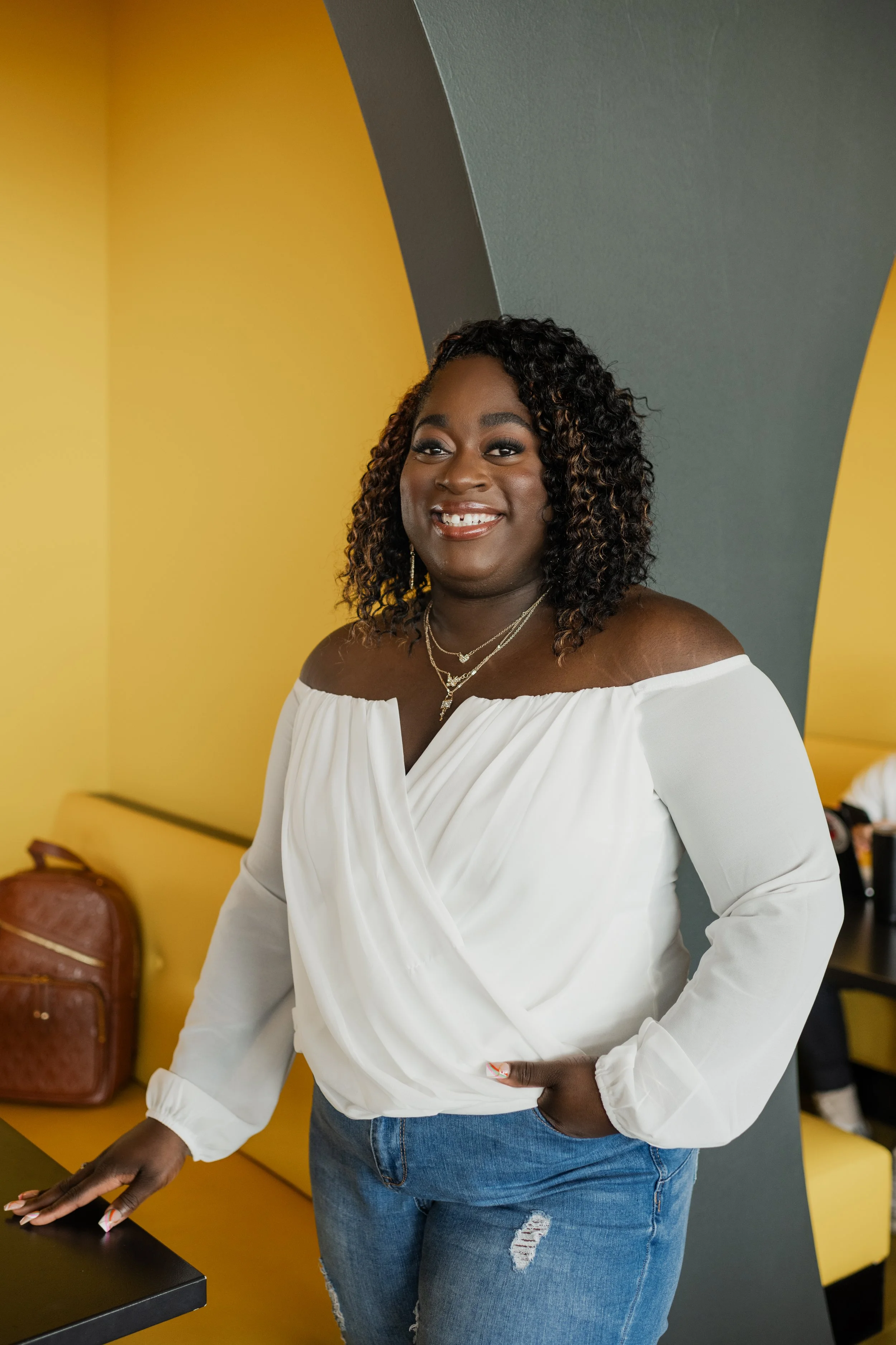 A woman with curly hair and a white off-the-shoulder blouse standing indoors, smiling, with yellow walls and a yellow couch in the background.