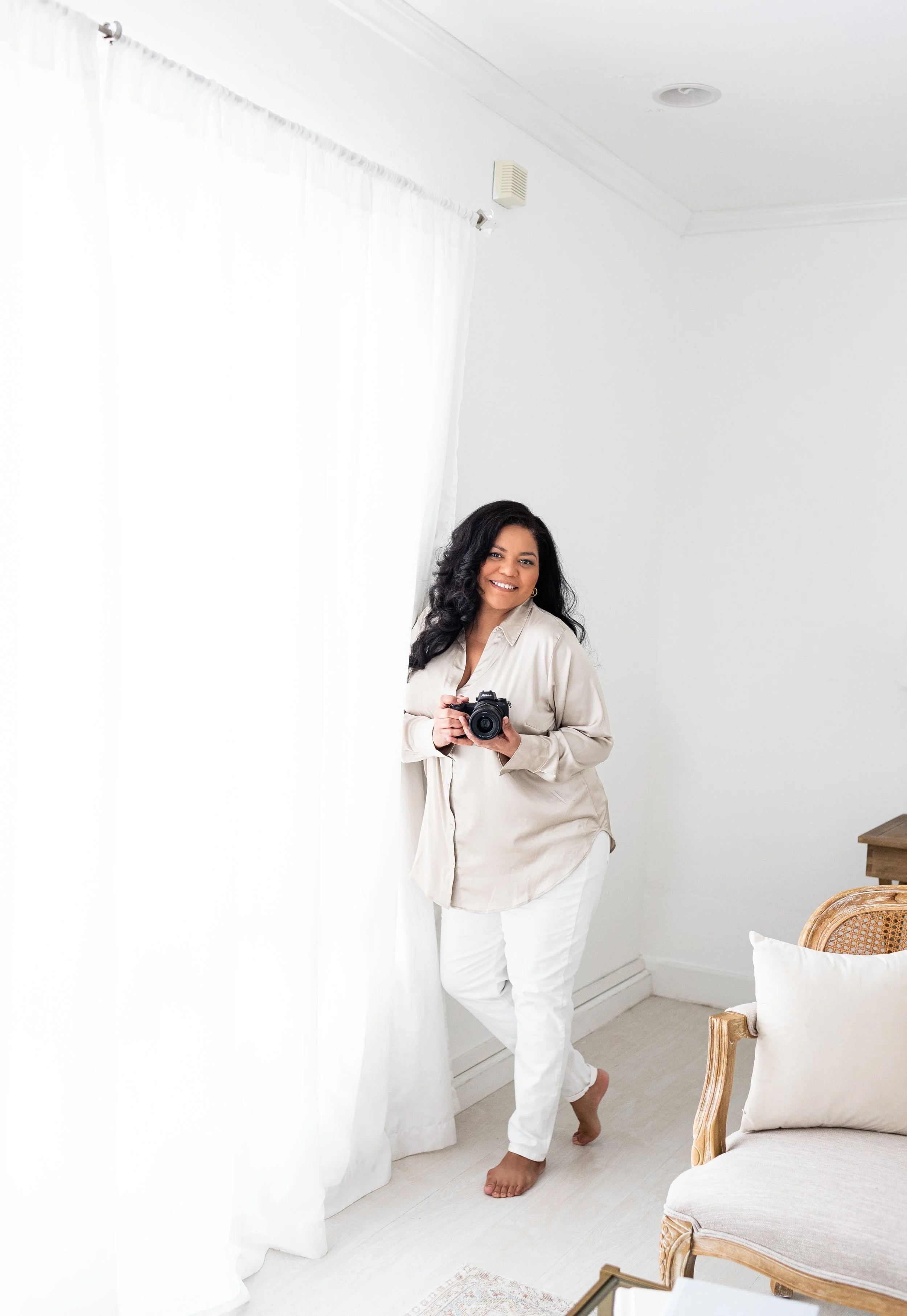 Woman standing barefoot in a bright room, holding a camera, smiling, near white curtains and light-colored furniture.
