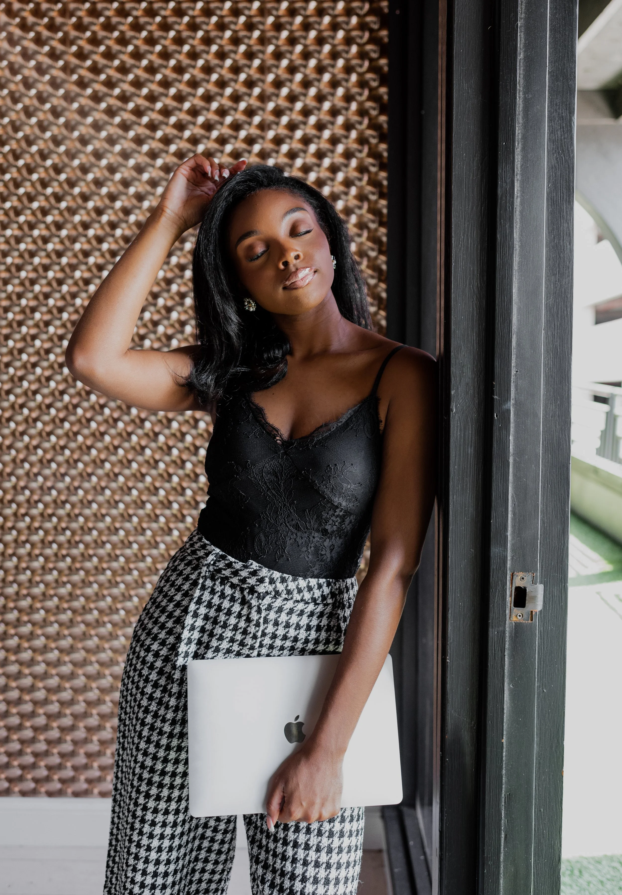 A woman with closed eyes, wearing a black lace camisole and houndstooth pants, holding a closed MacBook near her hip, standing by a dark wooden door with a textured wall in the background.
