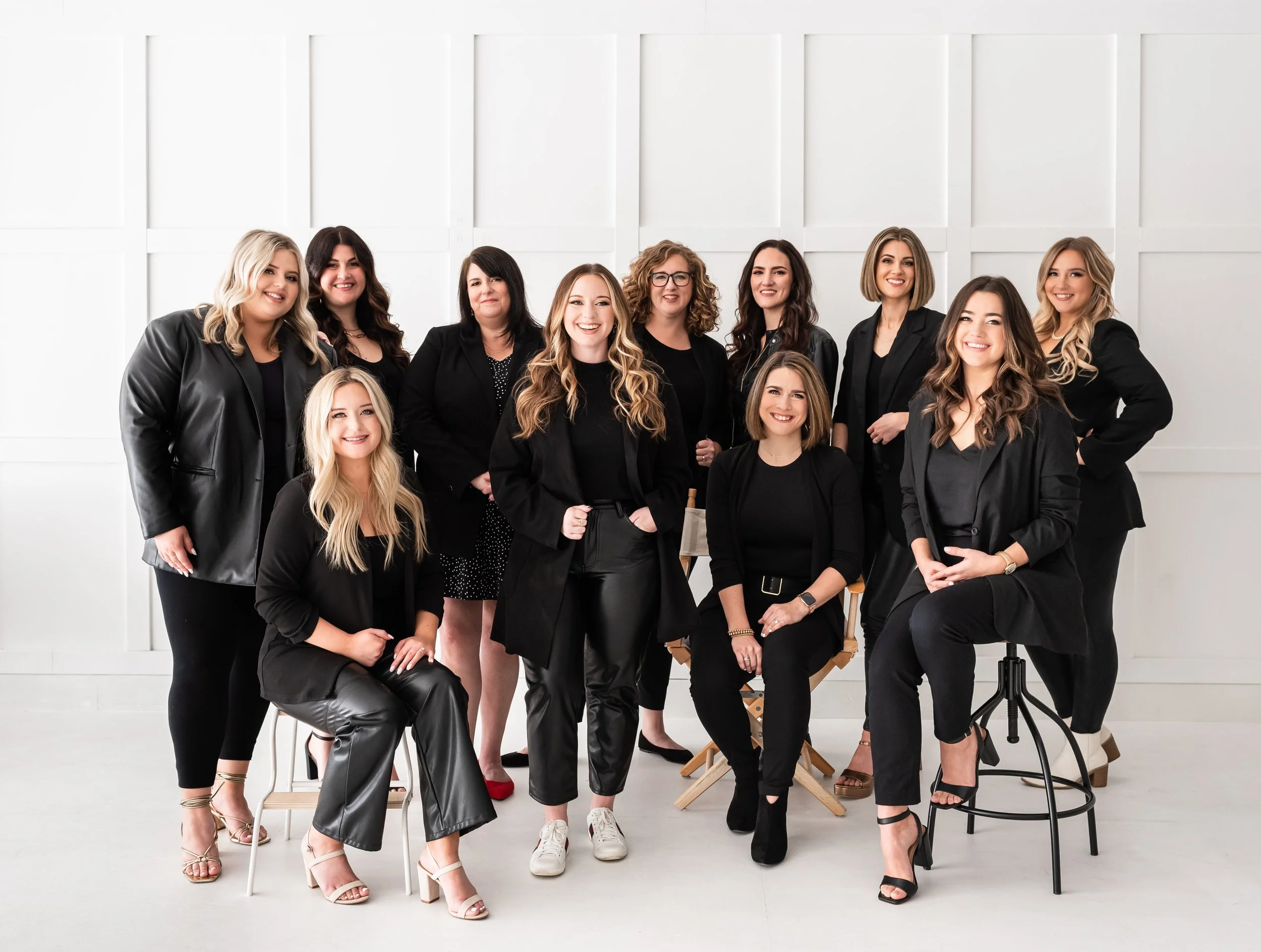 A diverse group of ten women dressed in black, posing together in a bright white studio with minimalist decor.