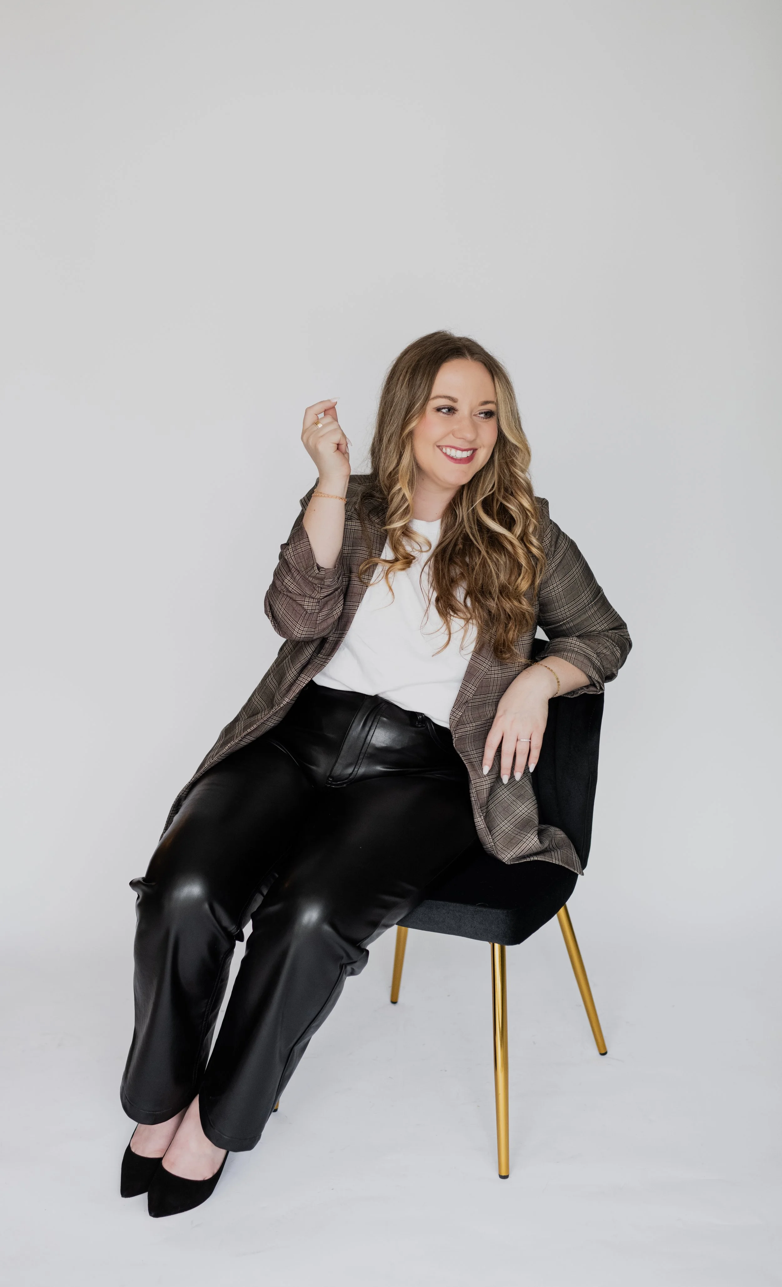 A woman with long wavy blonde hair, wearing a brown plaid blazer, white top, and black leather pants, sitting on a black chair with gold legs against a plain white background, smiling and looking to her left.