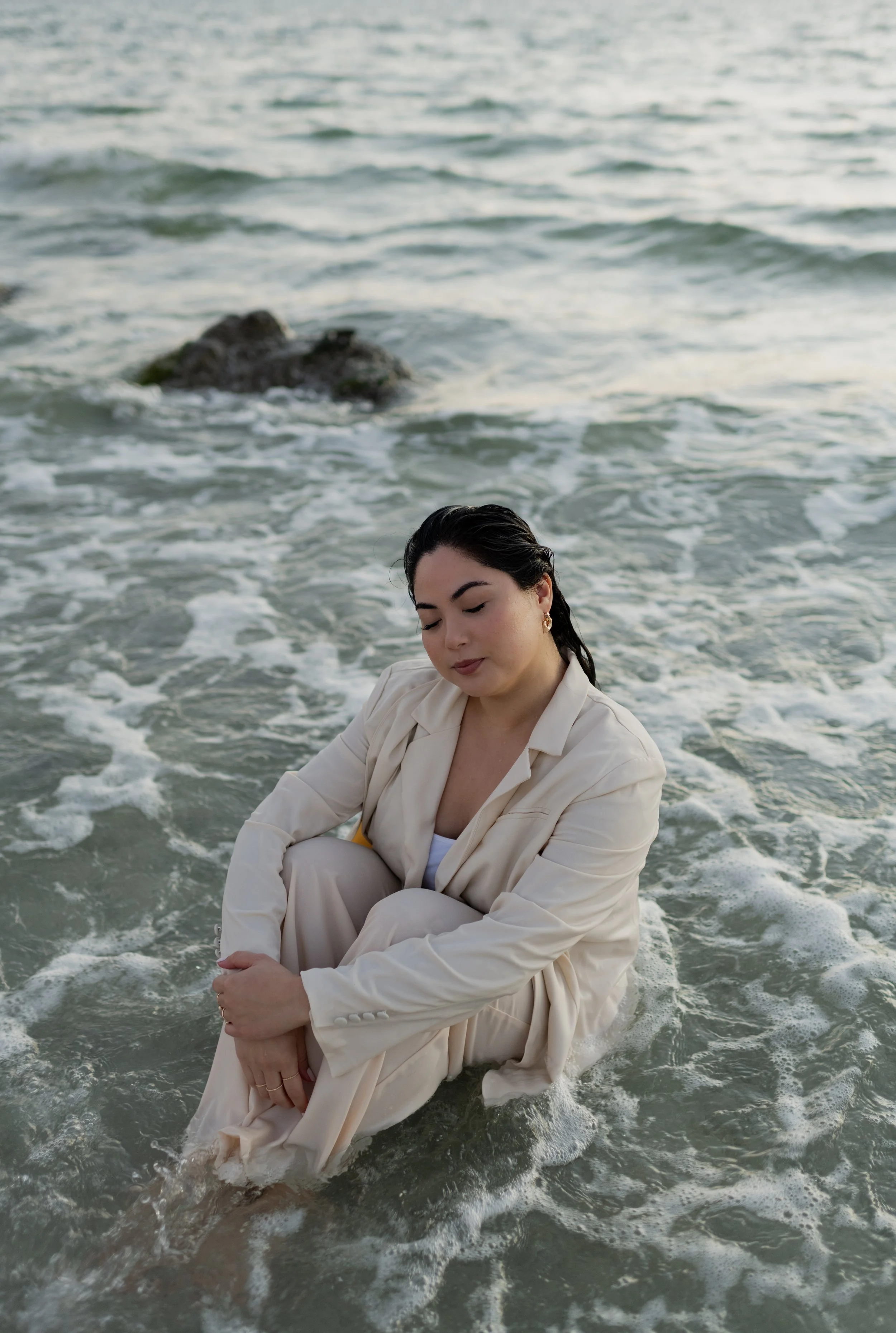 A woman in a cream-colored suit sitting in the water at the beach with her eyes closed and arms wrapped around her knees.