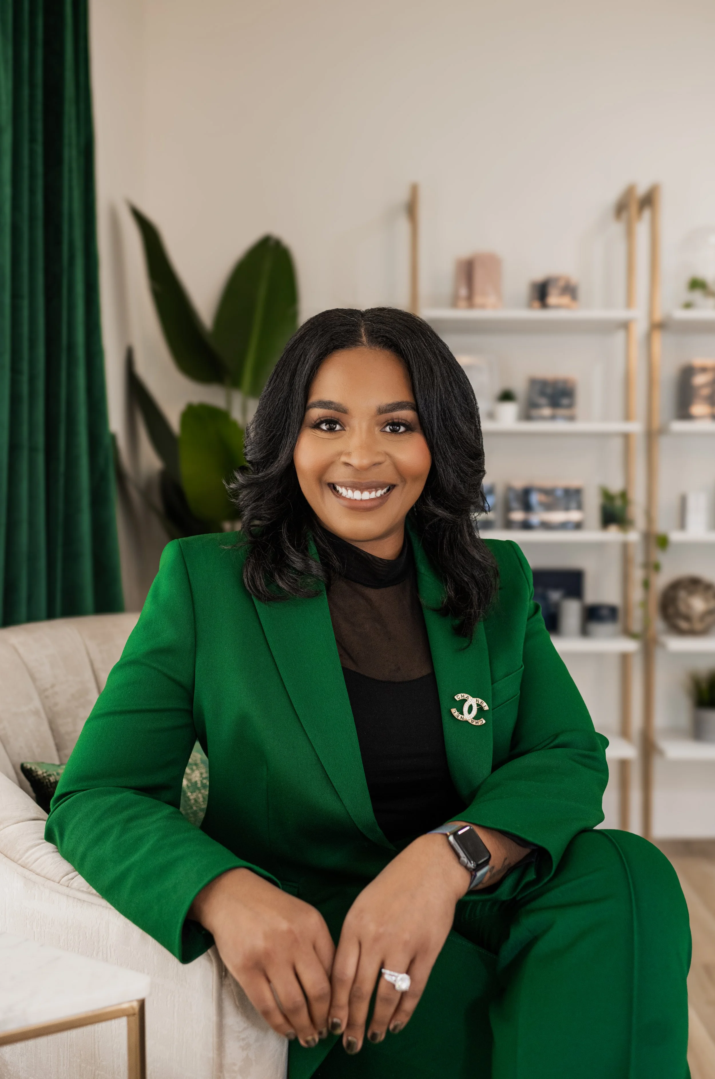 A woman with shoulder-length black hair wearing a bright green suit with a Chanel brooch, sitting on a cream-colored armchair in a modern living room.