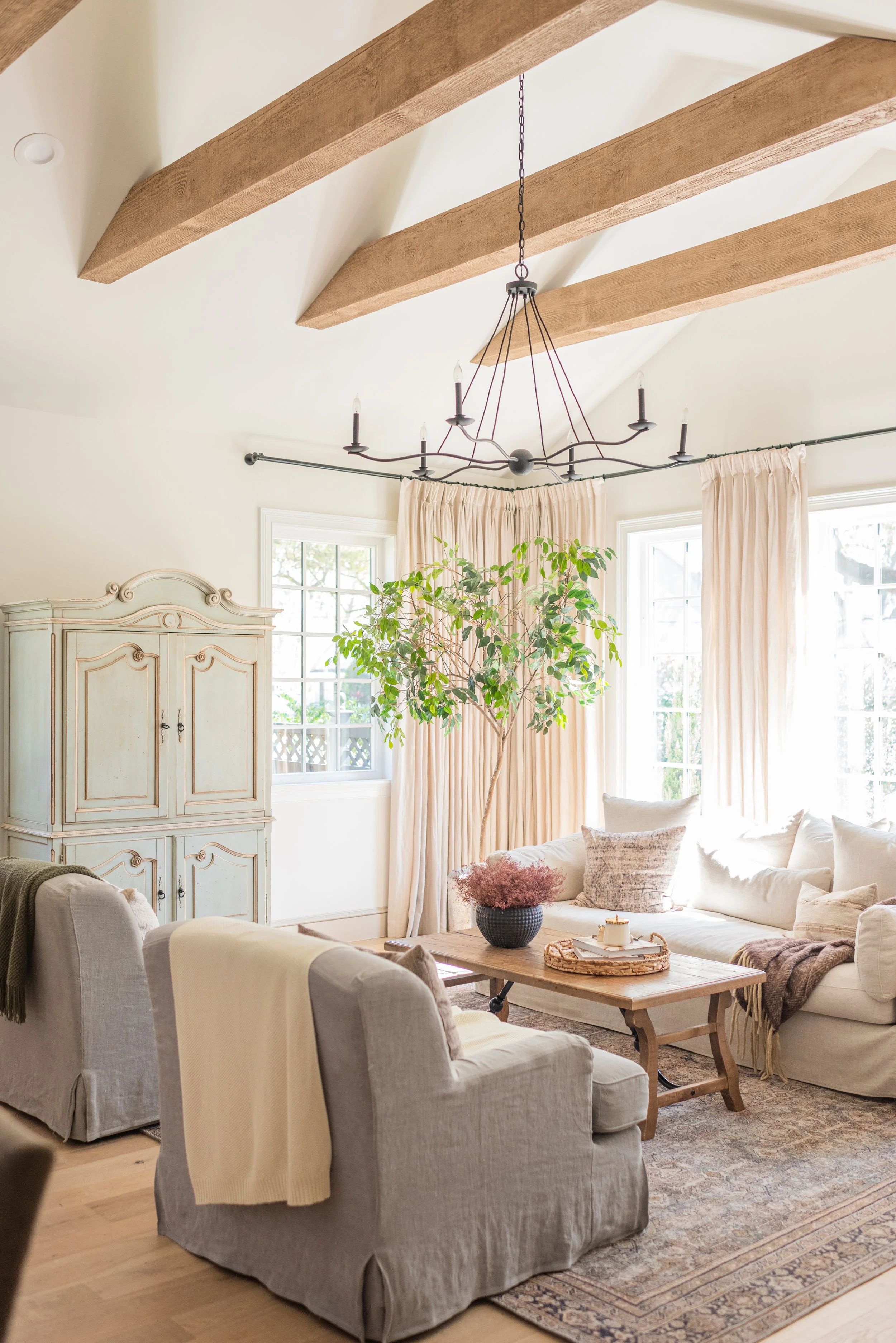 Bright living room with beige furniture, large windows with cream curtains, a vintage white armoire, a potted plant, and a rustic wooden coffee table with decorative items. Overhead, wooden beams and a black wrought-iron chandelier.
