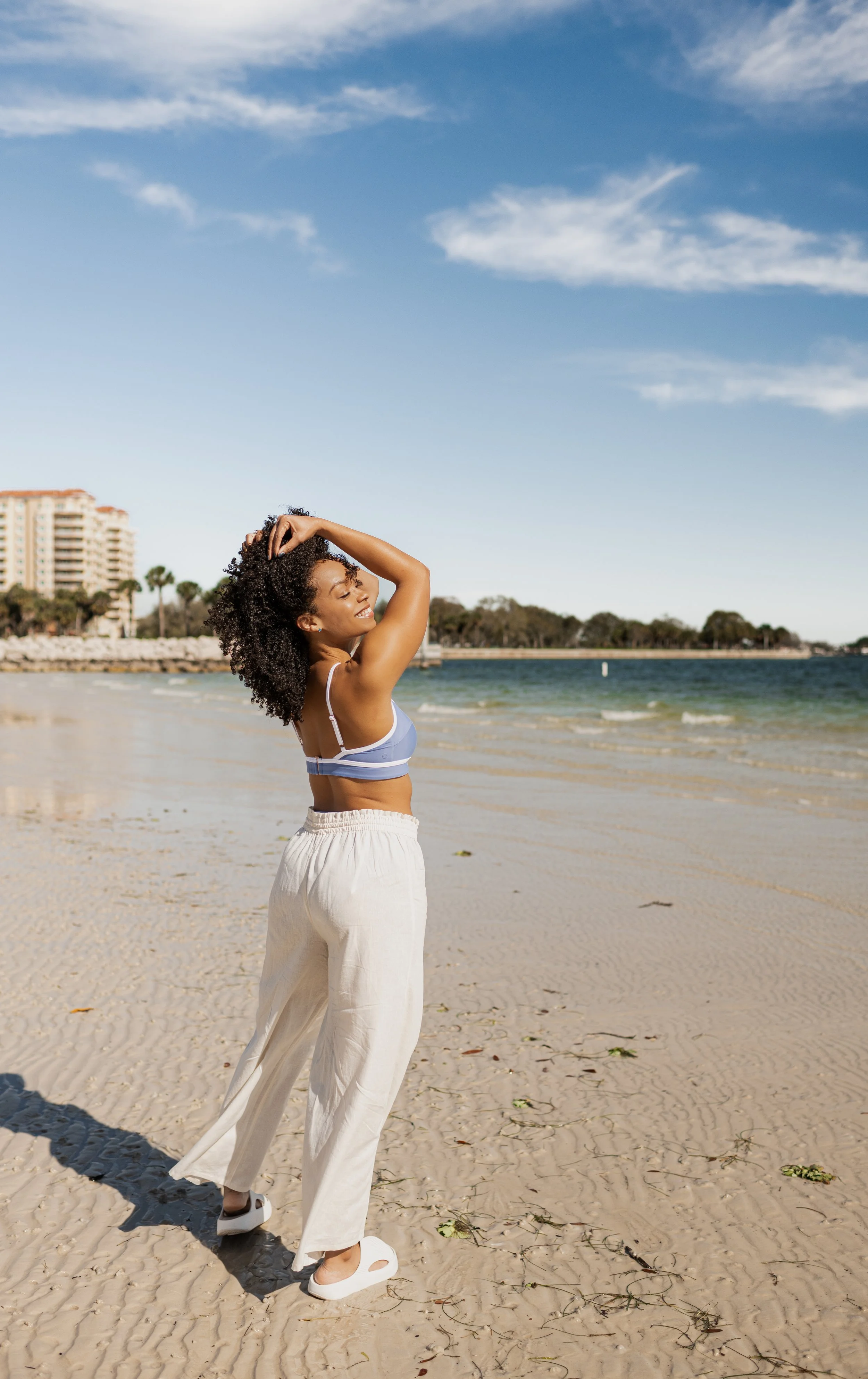 Woman in a striped bathing suit and white pants standing on sandy beach with calm water and buildings in the background under partly cloudy sky.
