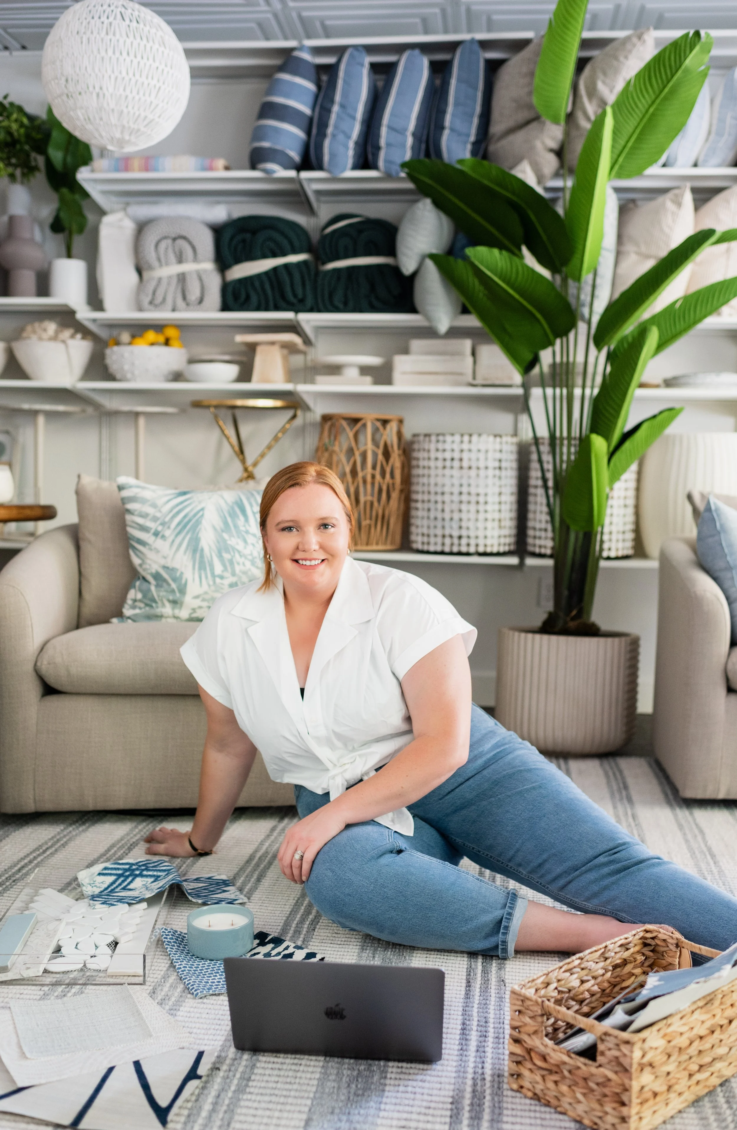 A woman is sitting on the floor in a living room, surrounded by textiles, fabric swatches, and a laptop, with a large green plant and shelves with rolled blankets and decorative objects behind her.