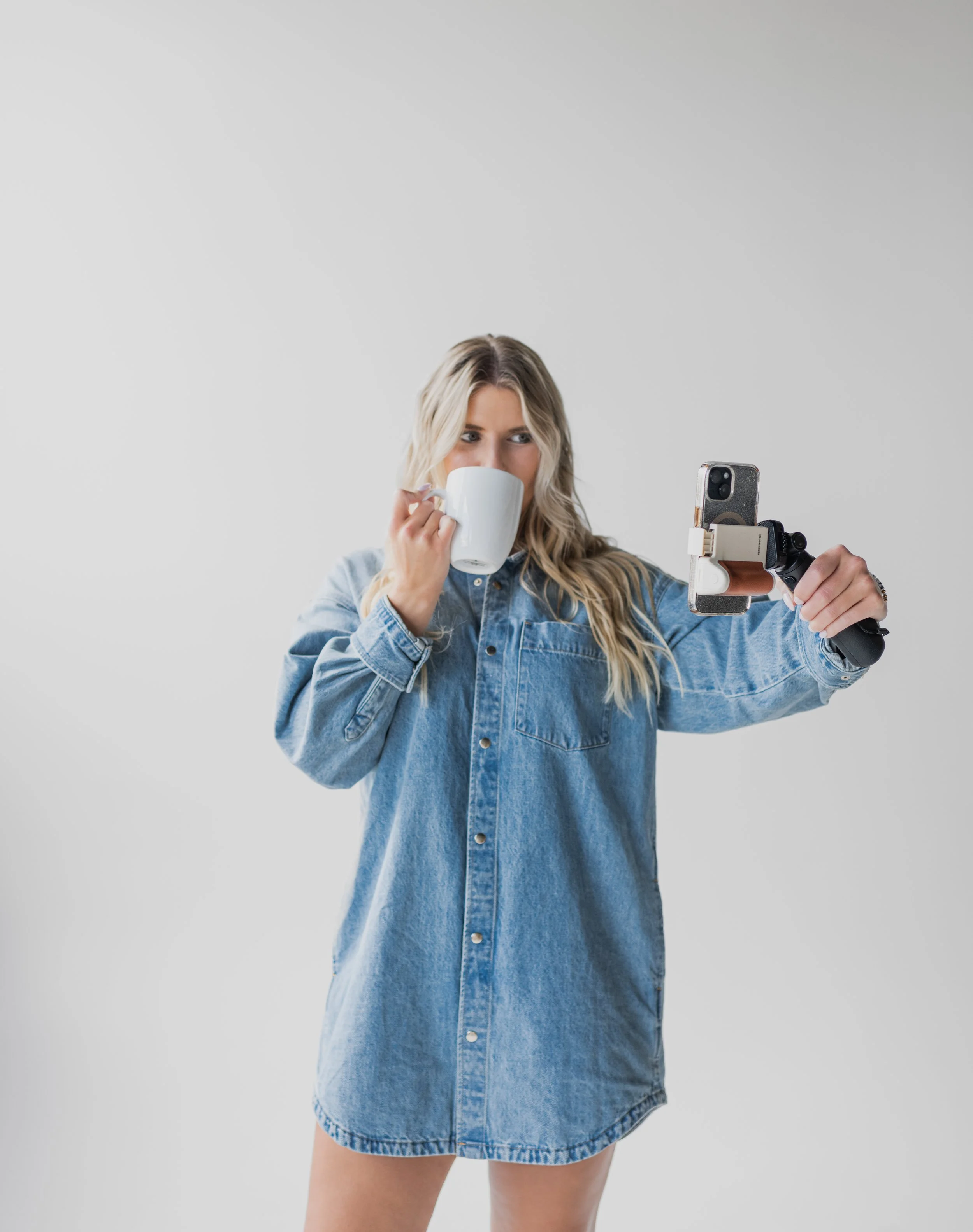 A woman with long blonde hair wearing a denim shirt dress, holding a smartphone in one hand and a coffee mug in the other, taking a selfie against a plain gray background.