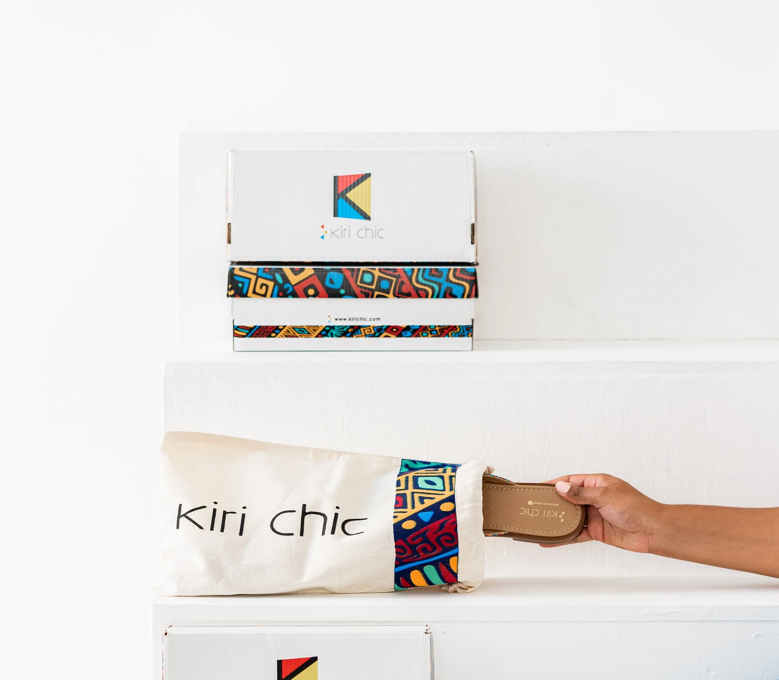 A person putting a flat shoe into a beige fabric bag with a colorful patterned stripe and the text "kiri chic" on it, with a stack of three colorful patterned boxes and a white box with a colorful logo behind it.