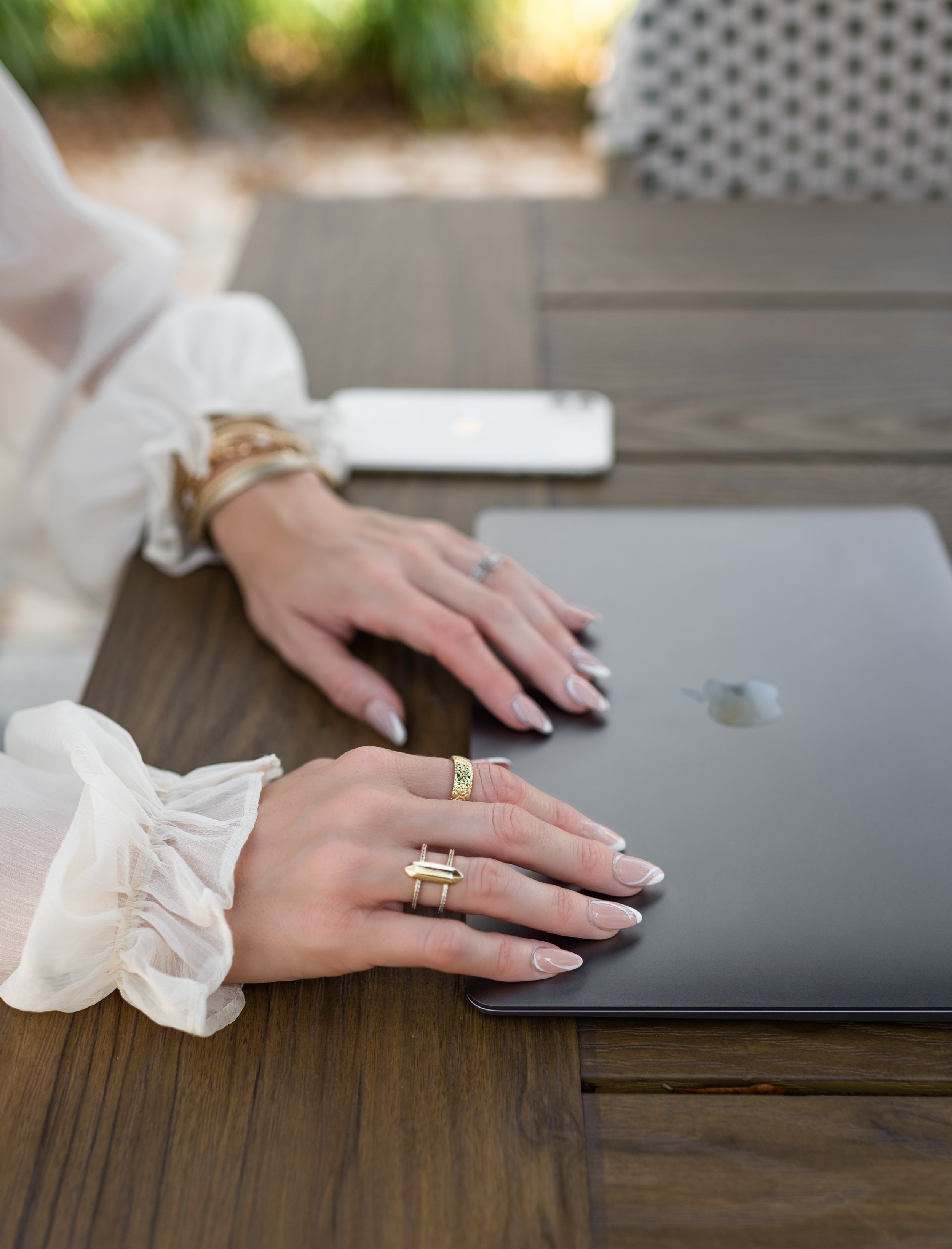 Person's hands with rings placed on a laptop on wooden table outdoors, with part of a tablet and a smartphone visible.
