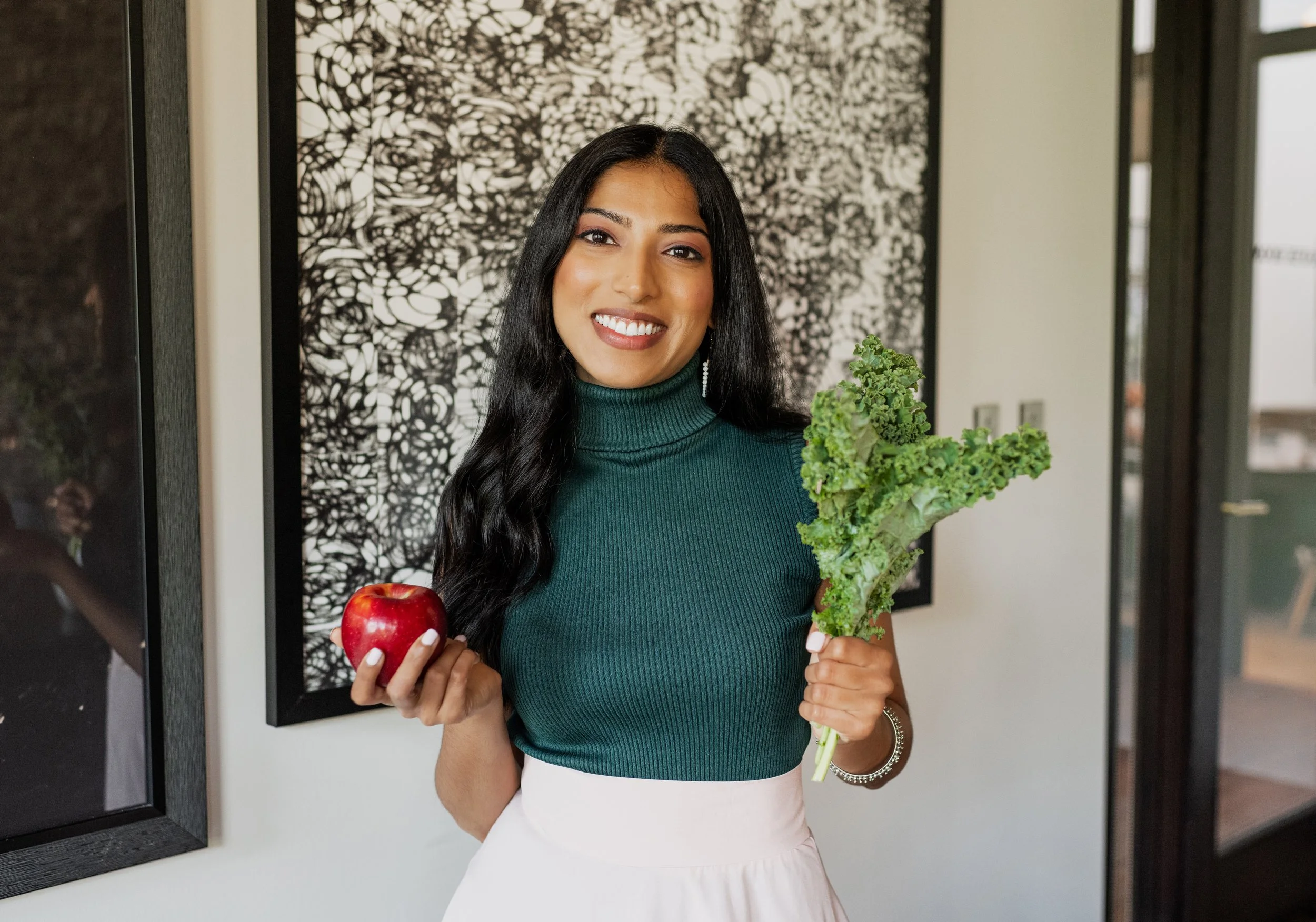 A young woman with long black hair smiling, holding a red apple in her right hand and a bunch of kale in her left, standing indoors in front of abstract wall art and windows.