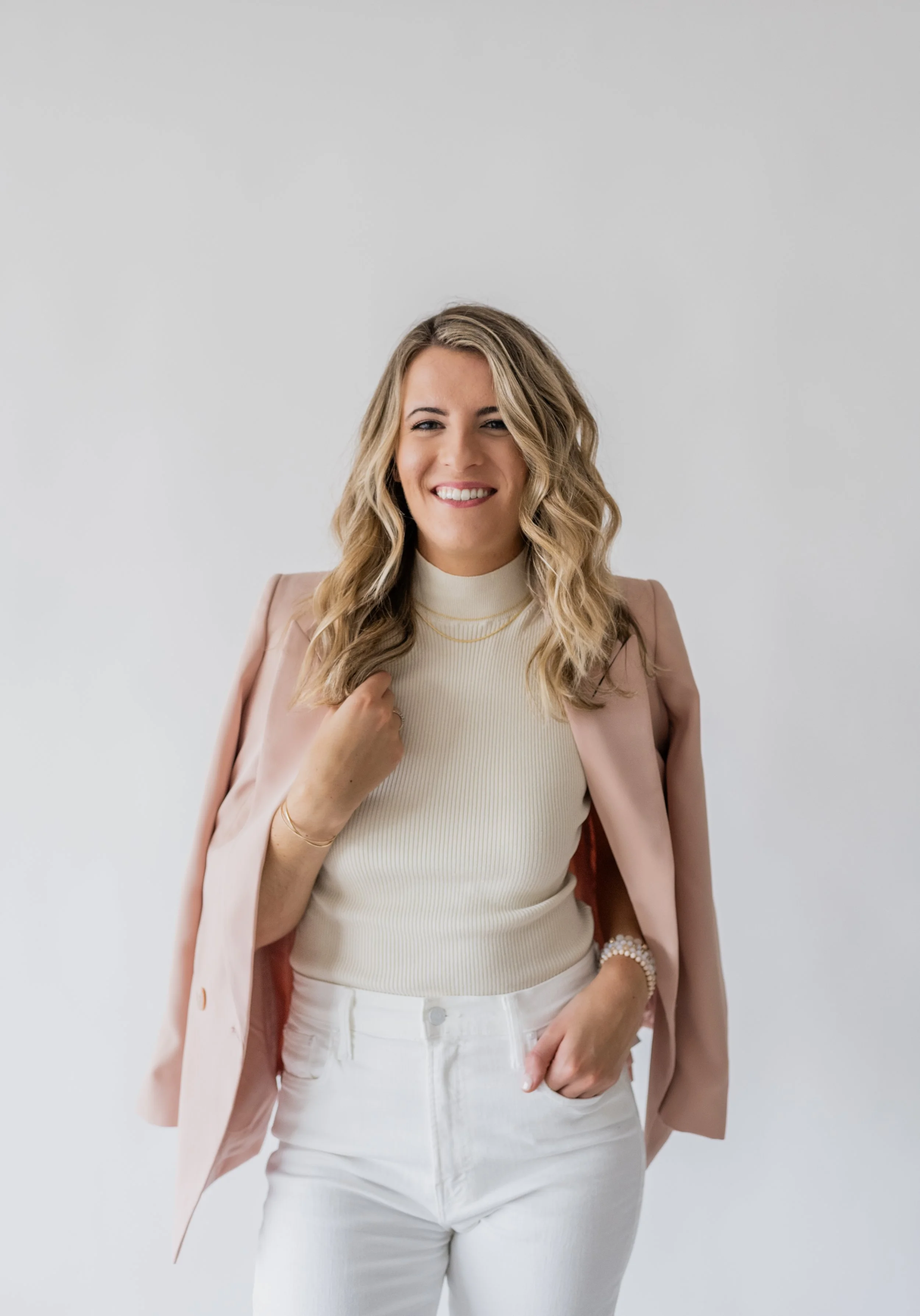 A smiling woman with wavy blonde hair wearing a beige turtleneck, white pants, and a pink blazer draped over her shoulders, standing against a plain light gray background.