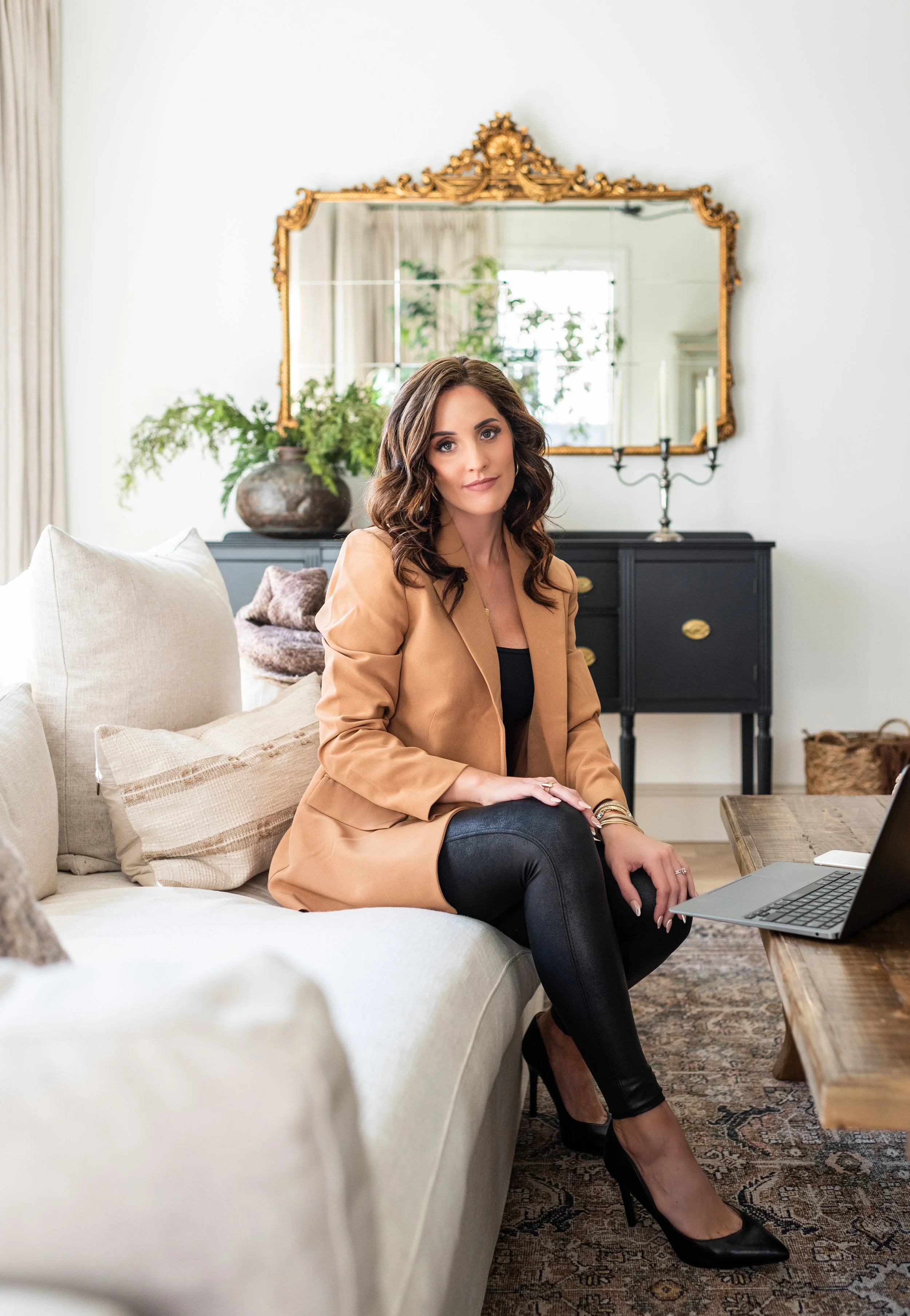 A woman sitting on a beige sofa in a living room, wearing a tan blazer, black top, black pants, and black high heels. There is a laptop on a wooden coffee table in front of her, and a large ornate mirror and decorative items in the background.