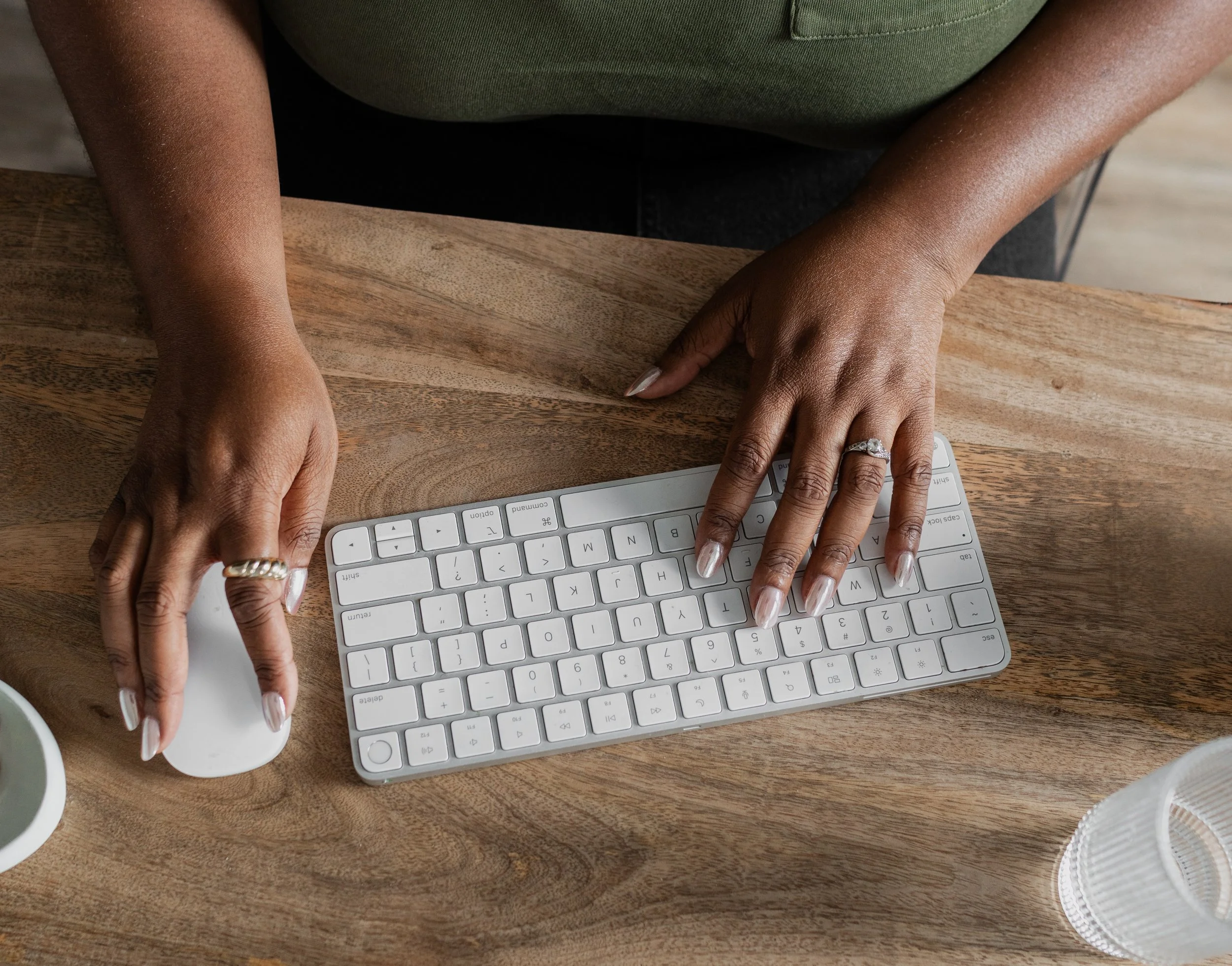 Person with dark skin using a white wireless keyboard and mouse on a wooden desk, with a cup of green beverage and a glass of water nearby