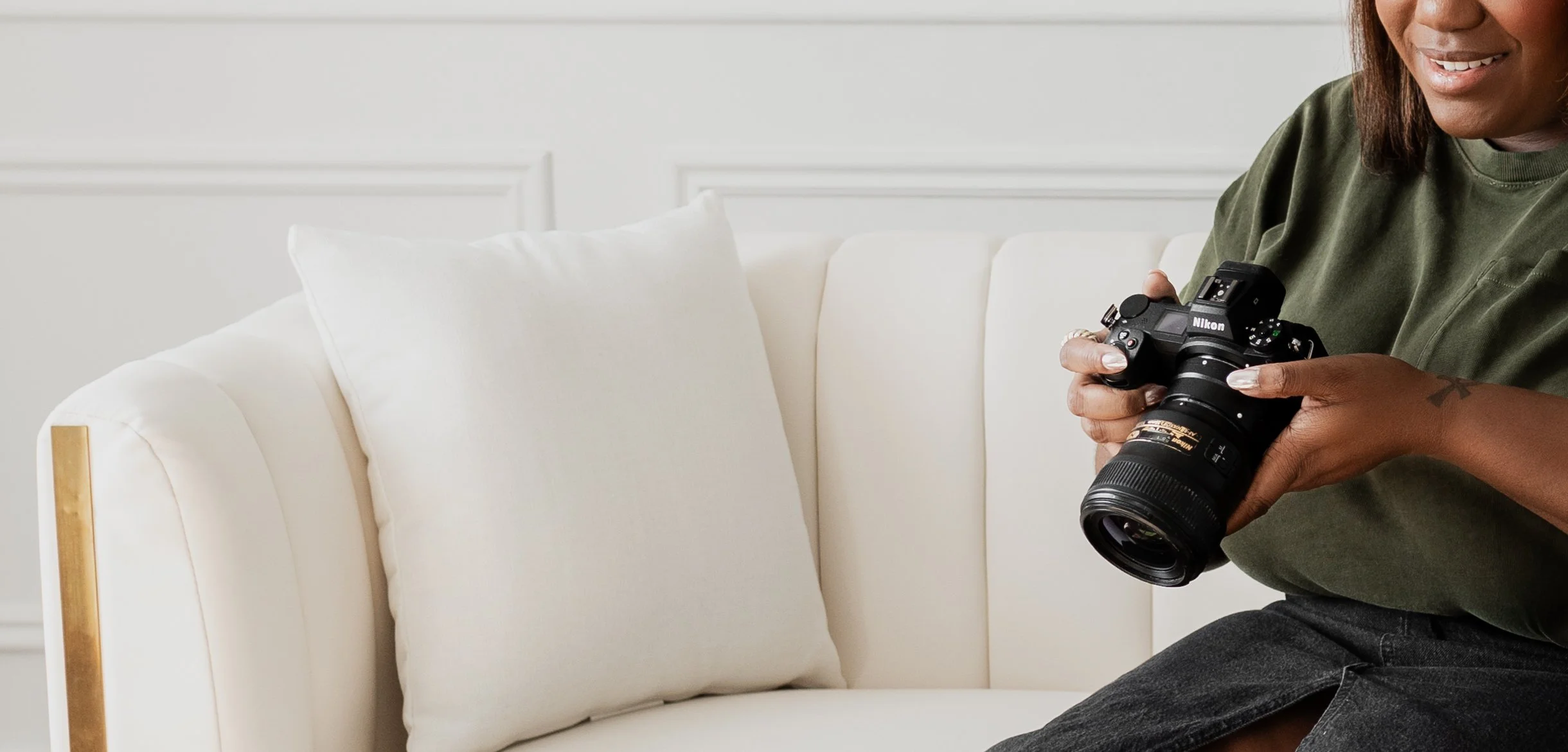 A person holding a Nikon DSLR camera, sitting on a white sofa with white pillows in a bright, minimalist room.