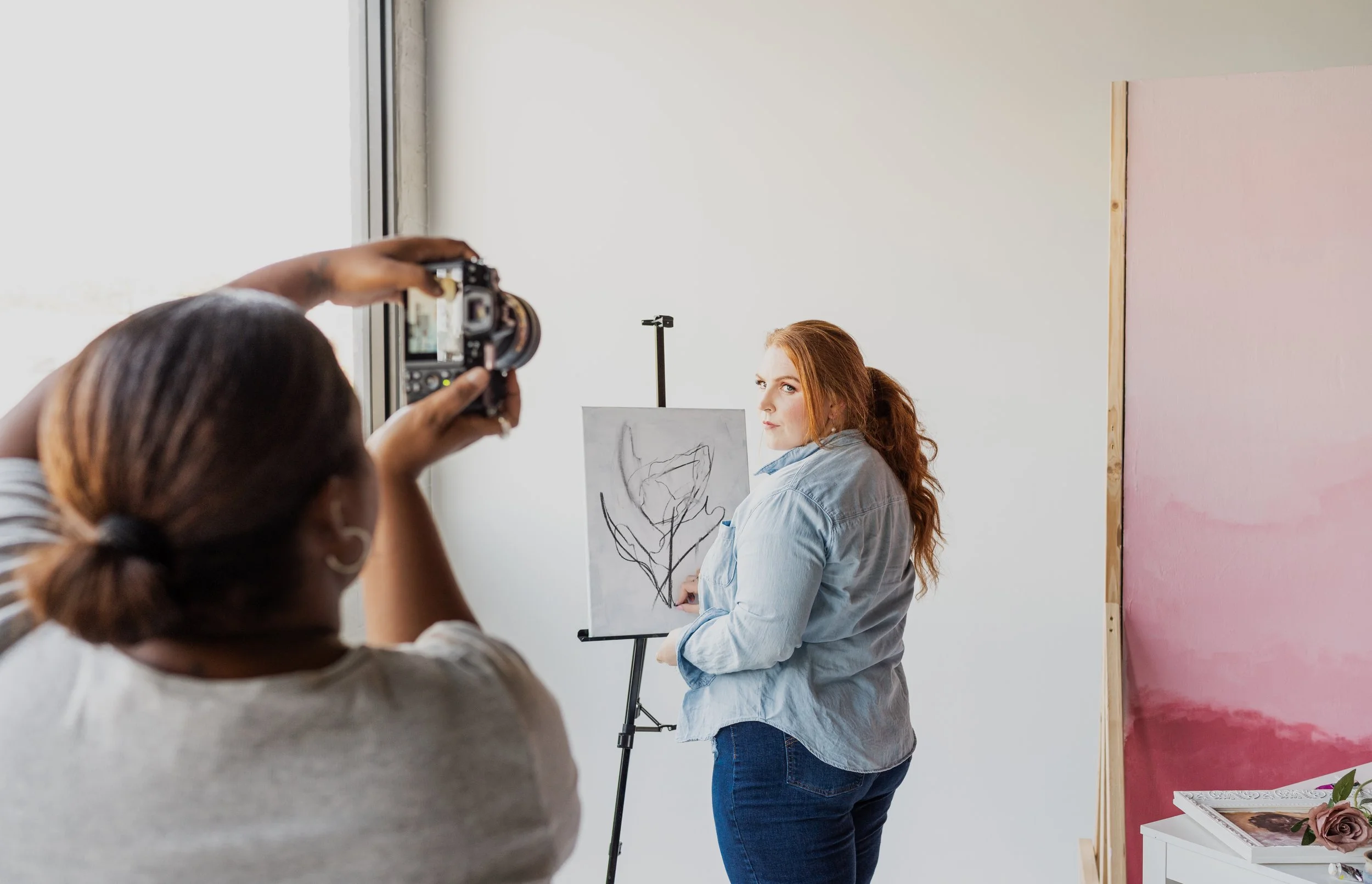 A woman with red hair, wearing a blue shirt, is posing next to a sketch of a flower on an easel for a photo being taken by another woman in a gray shirt, with a large pink artwork partially visible in the background.