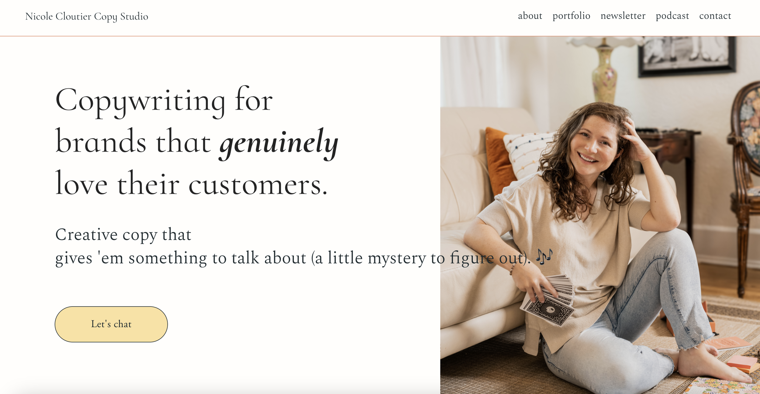 A woman with curly brown hair smiling and sitting on the floor by a sofa, holding tarot cards in one hand and touching her hair with the other. She is wearing a beige shirt and light blue jeans. The room is decorated with vintage furniture, a lamp, and framed artwork.