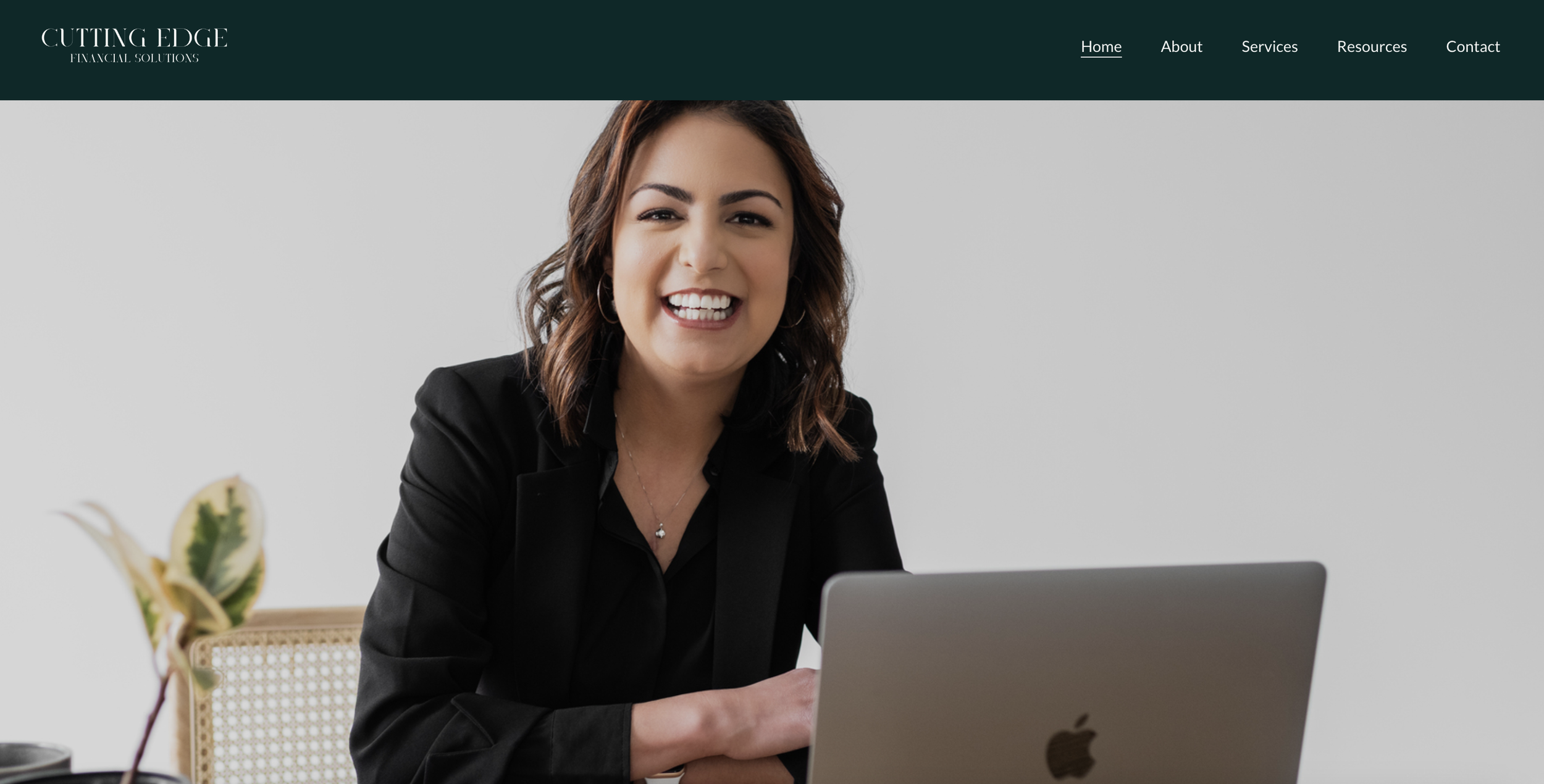 A woman with shoulder-length brown hair, smiling and wearing a black blazer, sitting at a desk with a silver MacBook laptop, in a professional setting.