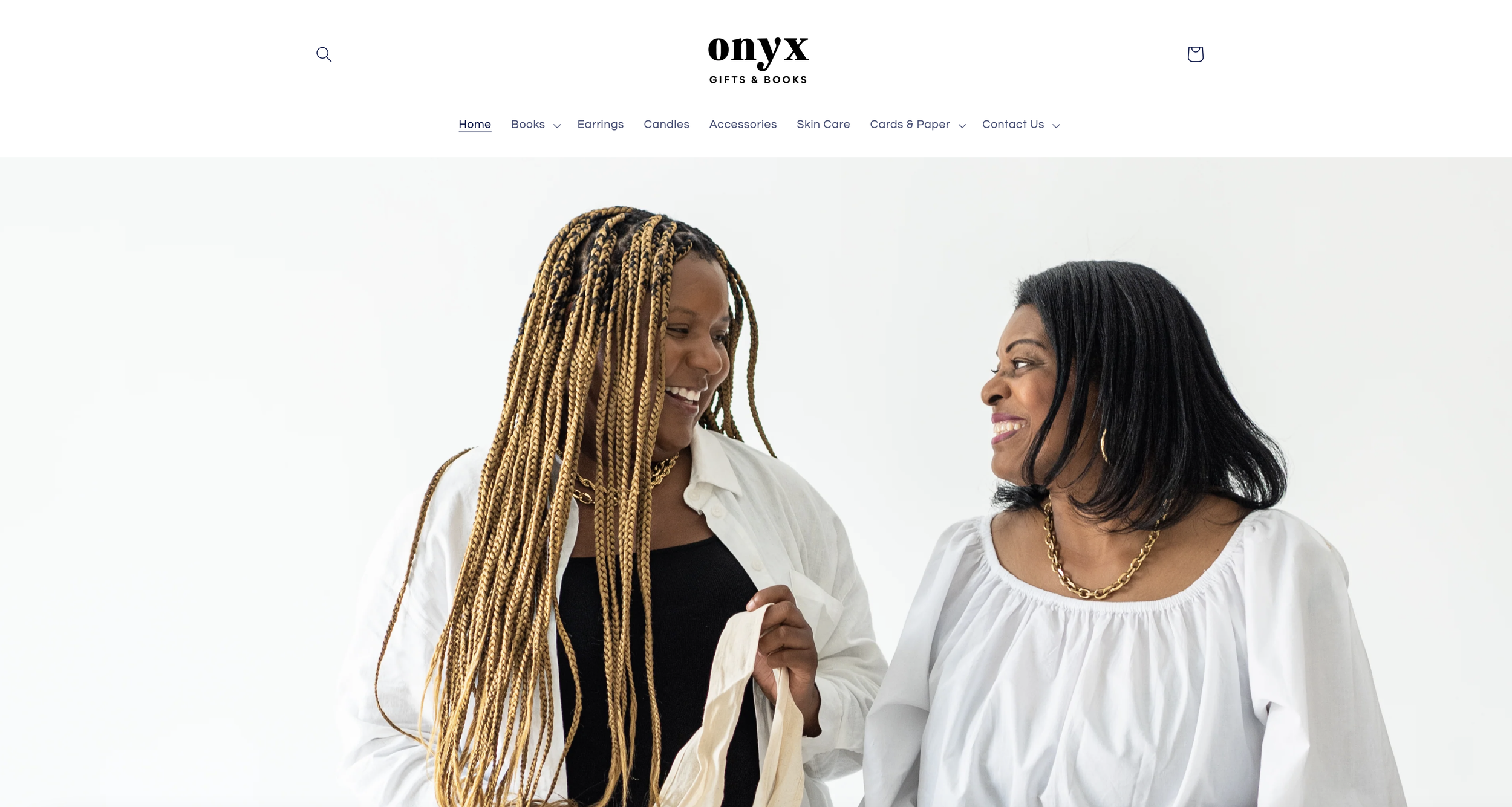 Two women smiling and talking to each other, one with long braided hair and the other with shoulder-length black hair, both wearing white tops, in front of a plain white background.