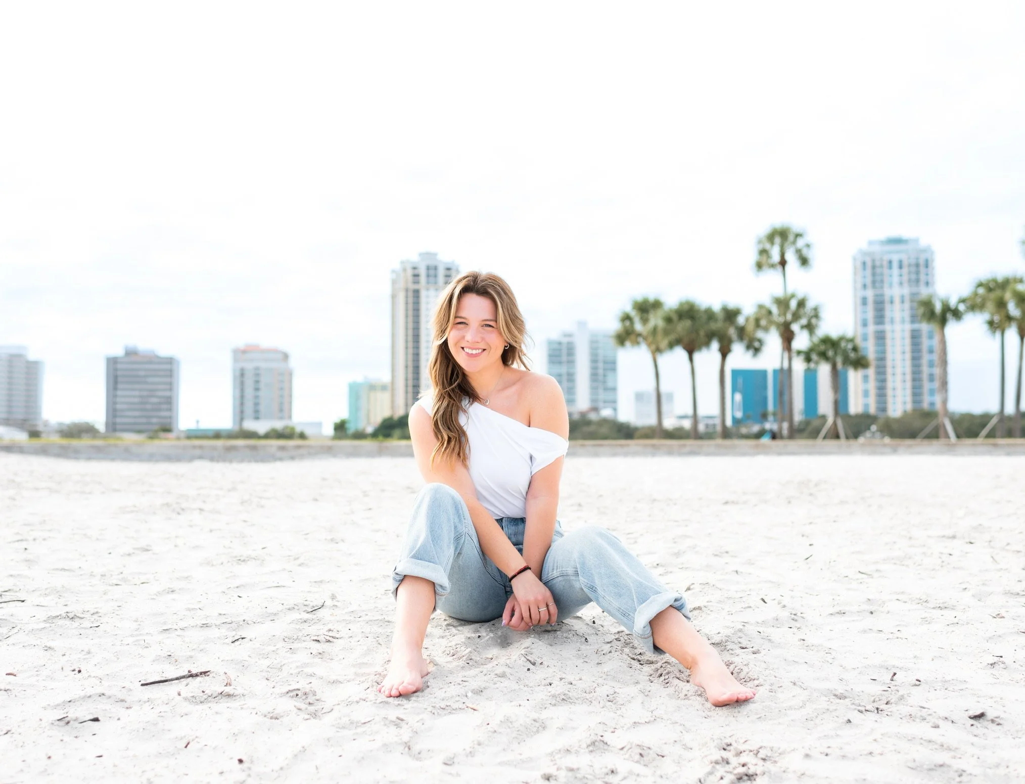 A smiling young woman sitting on a sandy beach with city buildings and palm trees in the background.