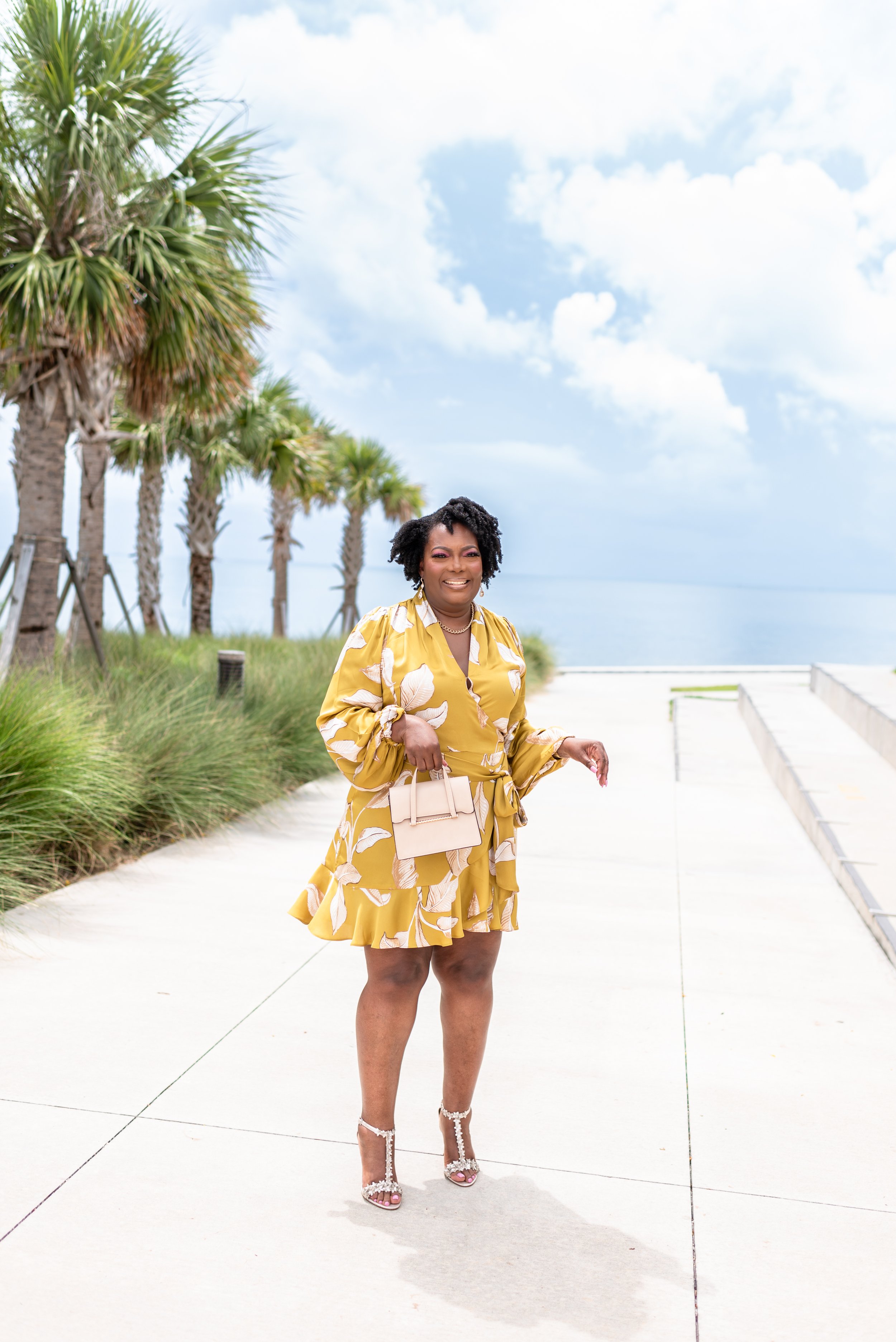 Woman in yellow floral dress holding a beige handbag, standing on a sunny pathway near palm trees by the ocean.