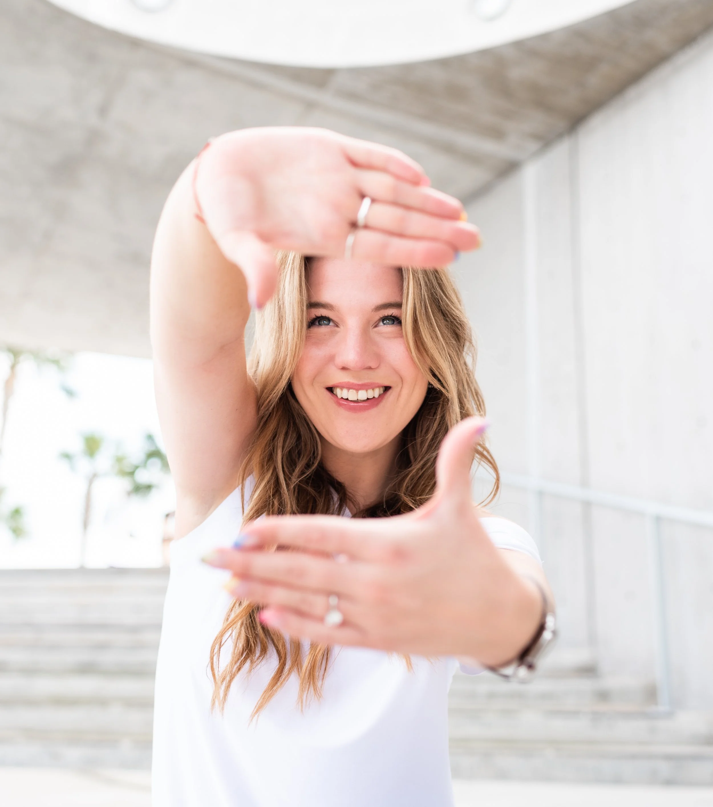 A woman with wavy blonde hair smiling as she frames her face with her hands in an outdoor urban setting.