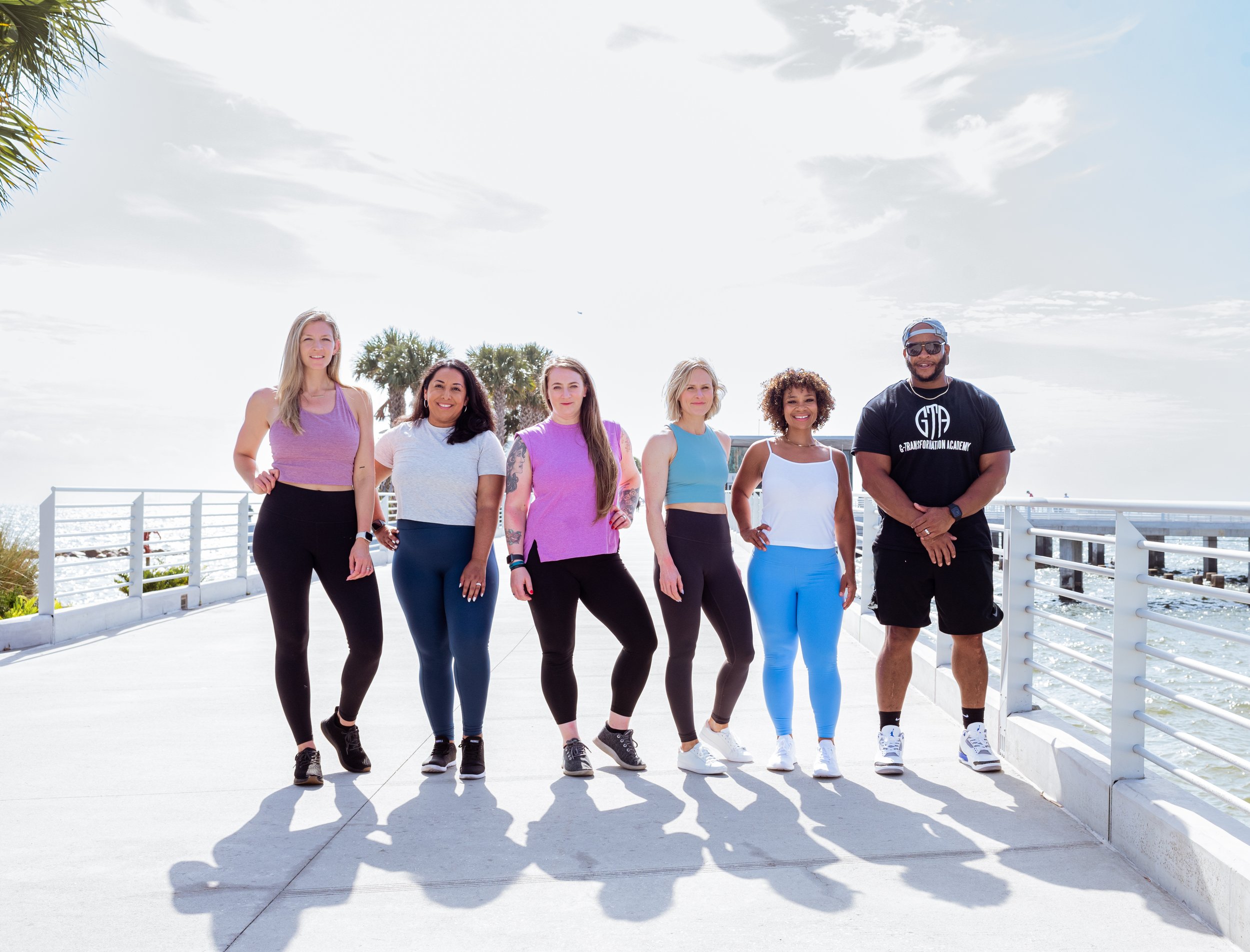 Group of six diverse young adults standing on a pier near the ocean, dressed in athletic wear, smiling, with palm trees and bright sky in the background.