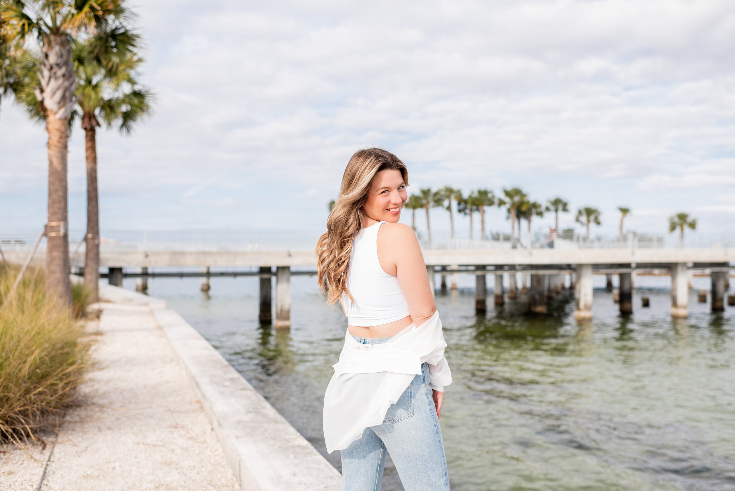 A woman with long wavy hair standing on a concrete path near water, smiling and looking over her shoulder, with palm trees and a pier in the background under a partly cloudy sky.