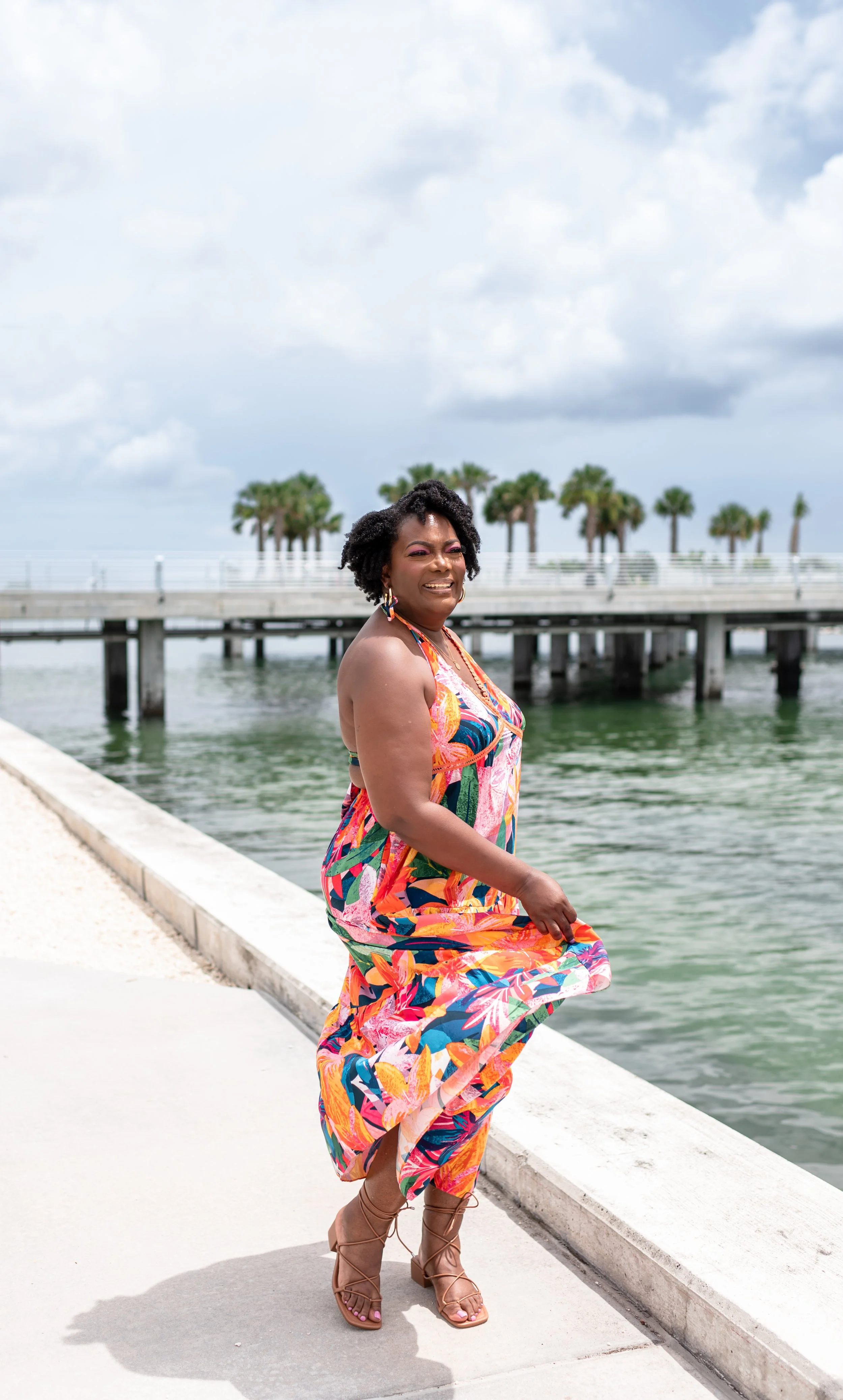 A woman in a colorful tropical dress and high-heeled sandals stands by a waterfront with a pier and palm trees in the background, smiling.