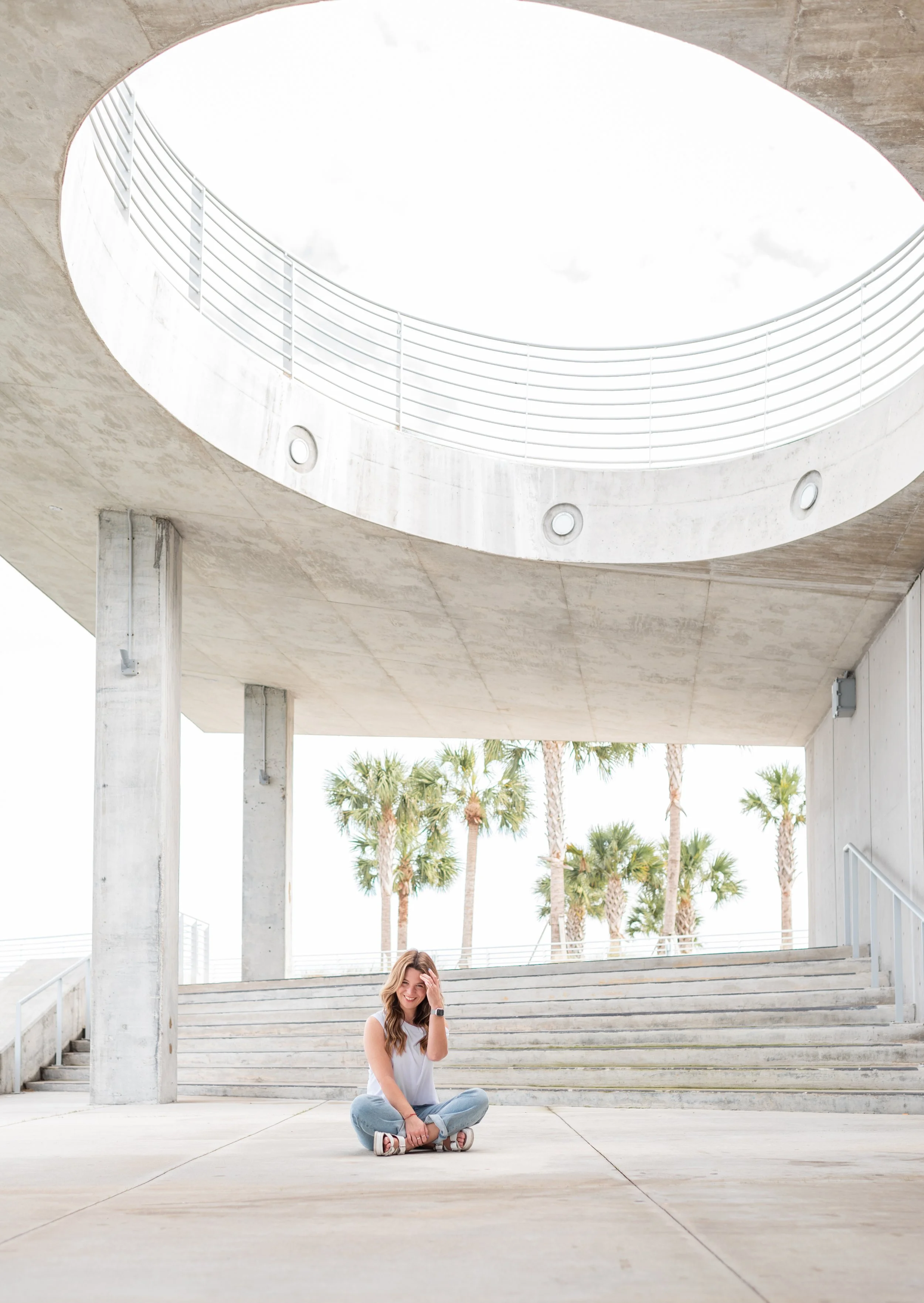 A smiling woman sitting cross-legged on the pavement under a modern concrete building with a circular opening above, and palm trees in the background.