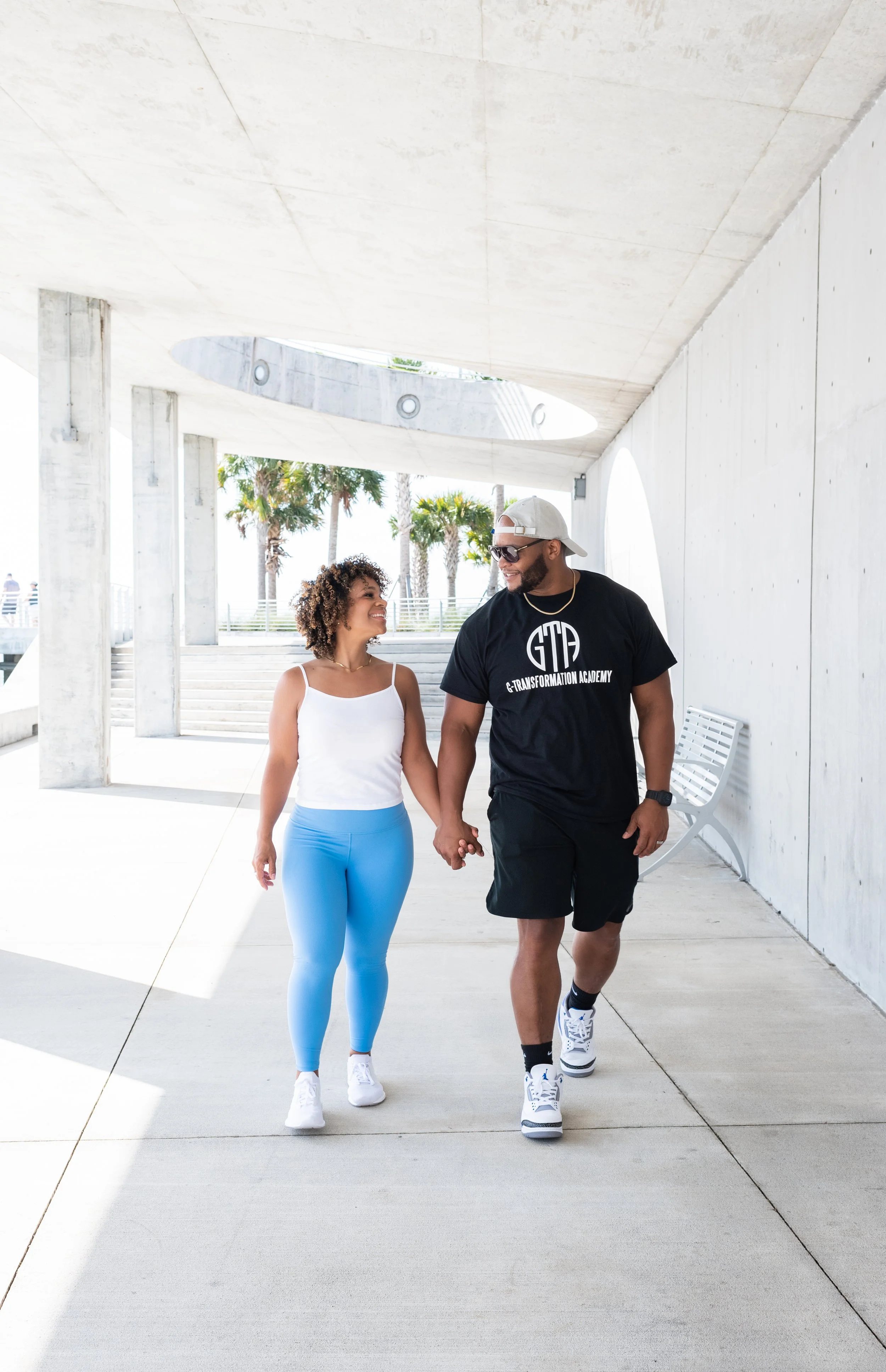 A couple walking hand in hand outdoors under a concrete structure with palm trees in the background, smiling at each other.