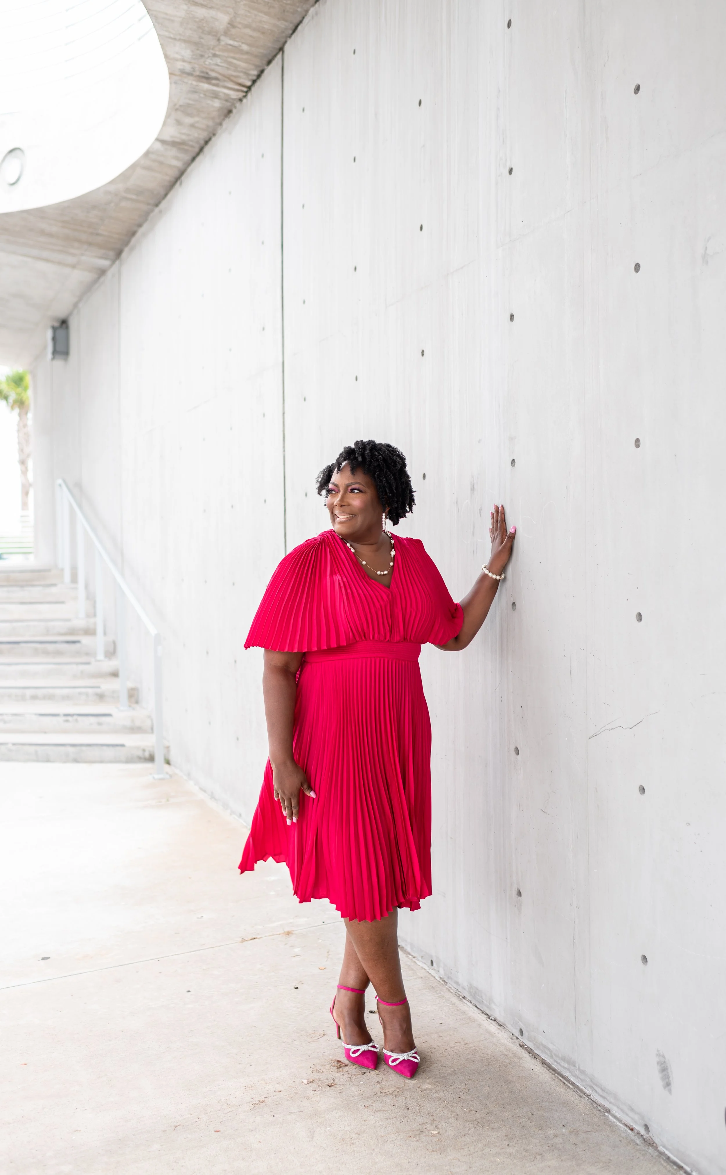 Woman in a bright pink pleated dress and matching heels posing against a white concrete wall.