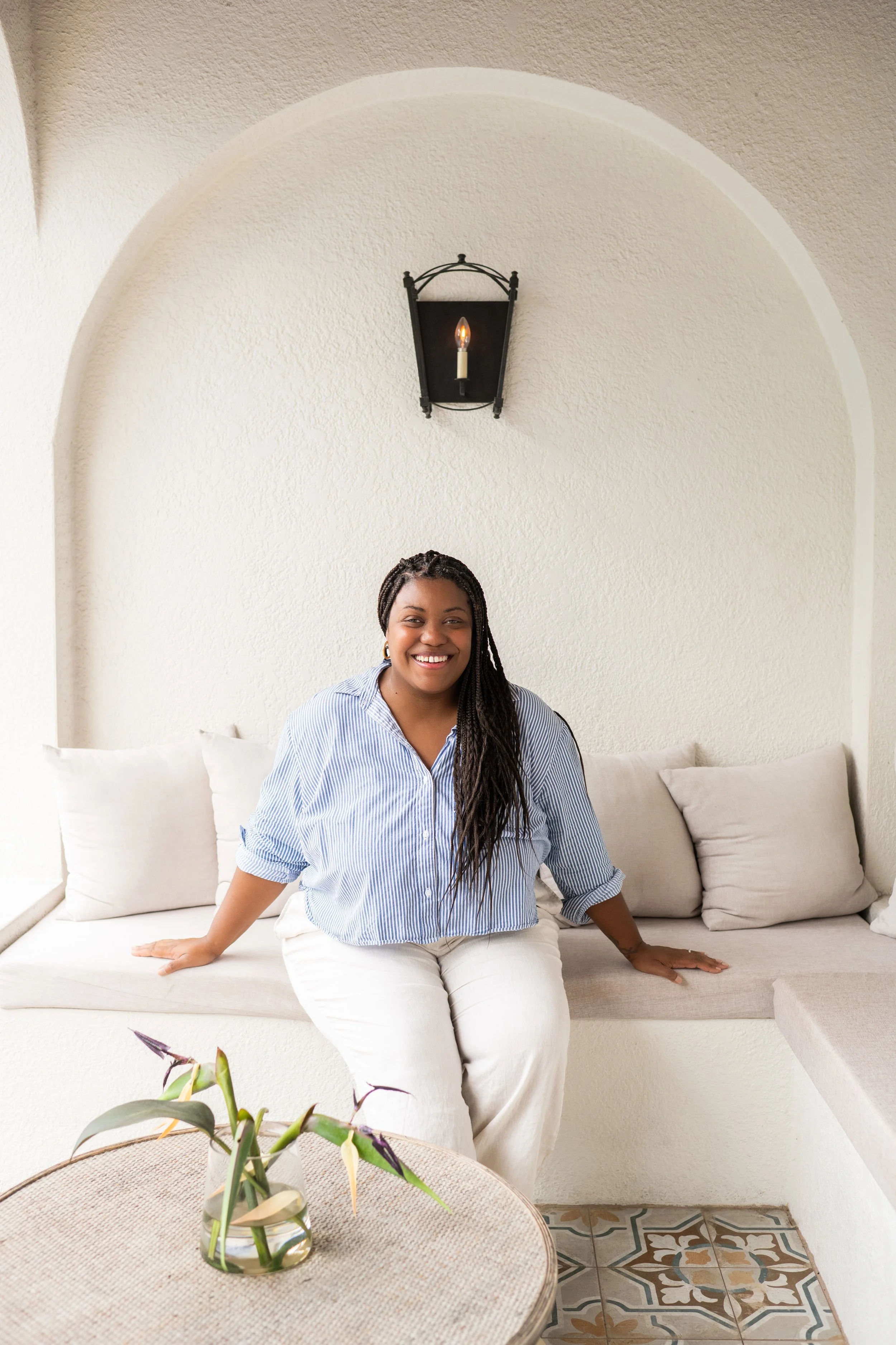A woman with long braided hair sitting on a built-in sofa in a room with white textured walls and a lantern-style light fixture. A small round table with a glass vase holding a plant is in the foreground.