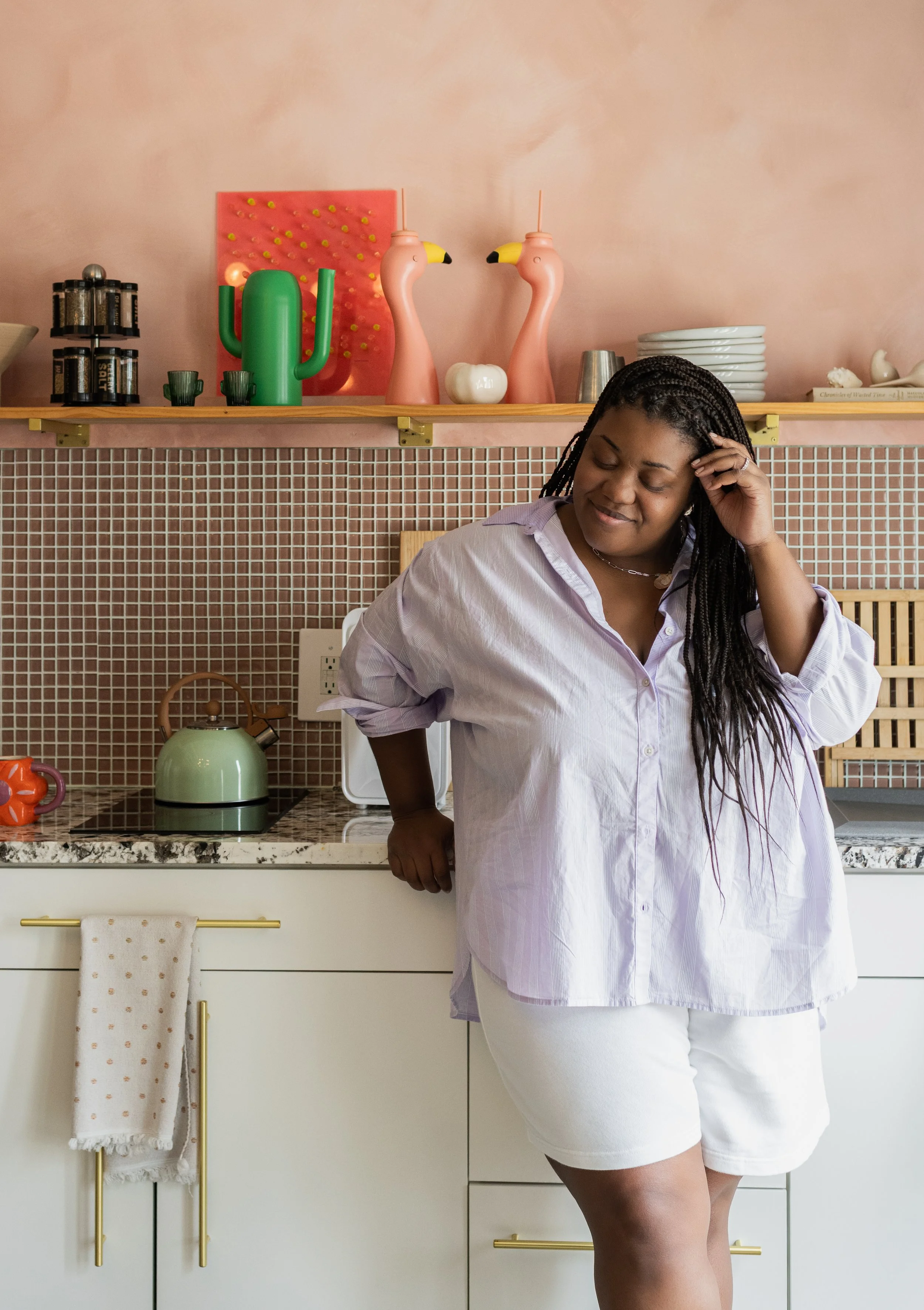 A woman in a white shirt and shorts standing in a modern kitchen with pink tiled backsplash, decorative flamingo and cactus decor on a shelf, and a cream-colored cabinet.