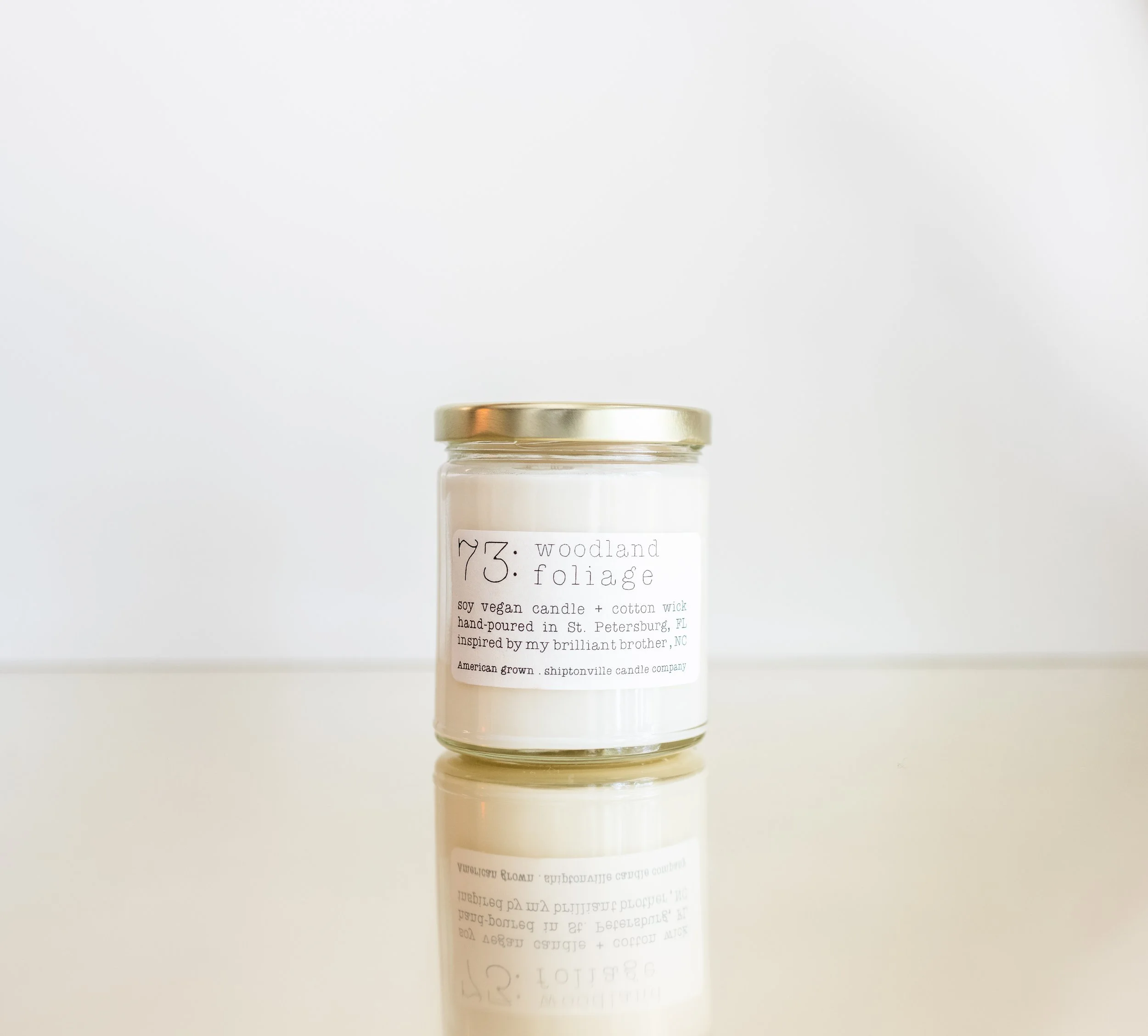 A glass jar with a gold lid containing a white candle labeled 'woodland foliage' soy vegan candle with cotton wick, hand-poured in St. Petersburg, Florida, inspired by the creator's brother, North Carolina, made by Shiptonville candle company, and re