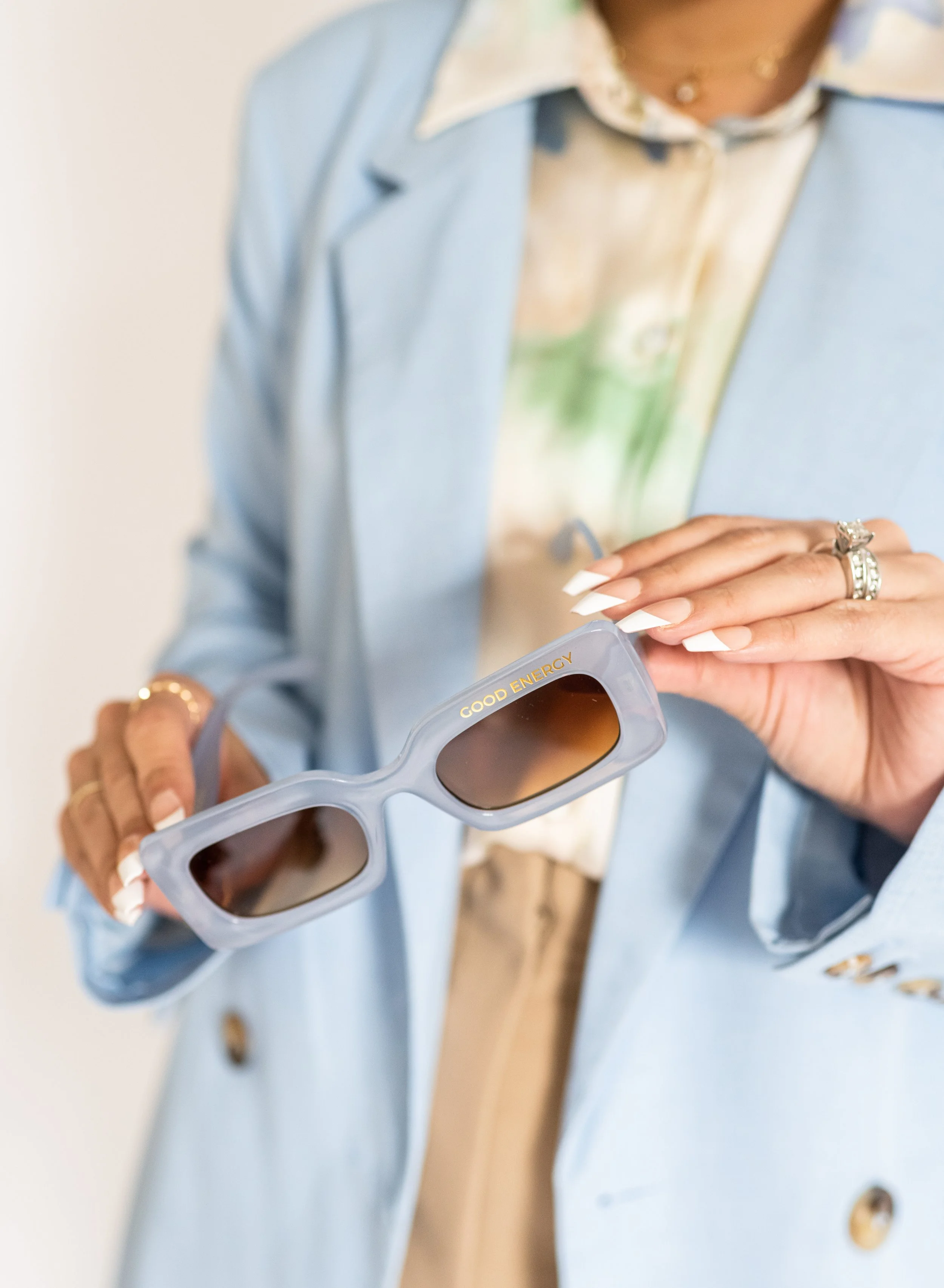Person holding sunglasses with gold letters 'Good Energy' on the side, wearing rings, a blue blazer, and a photo-printed shirt.