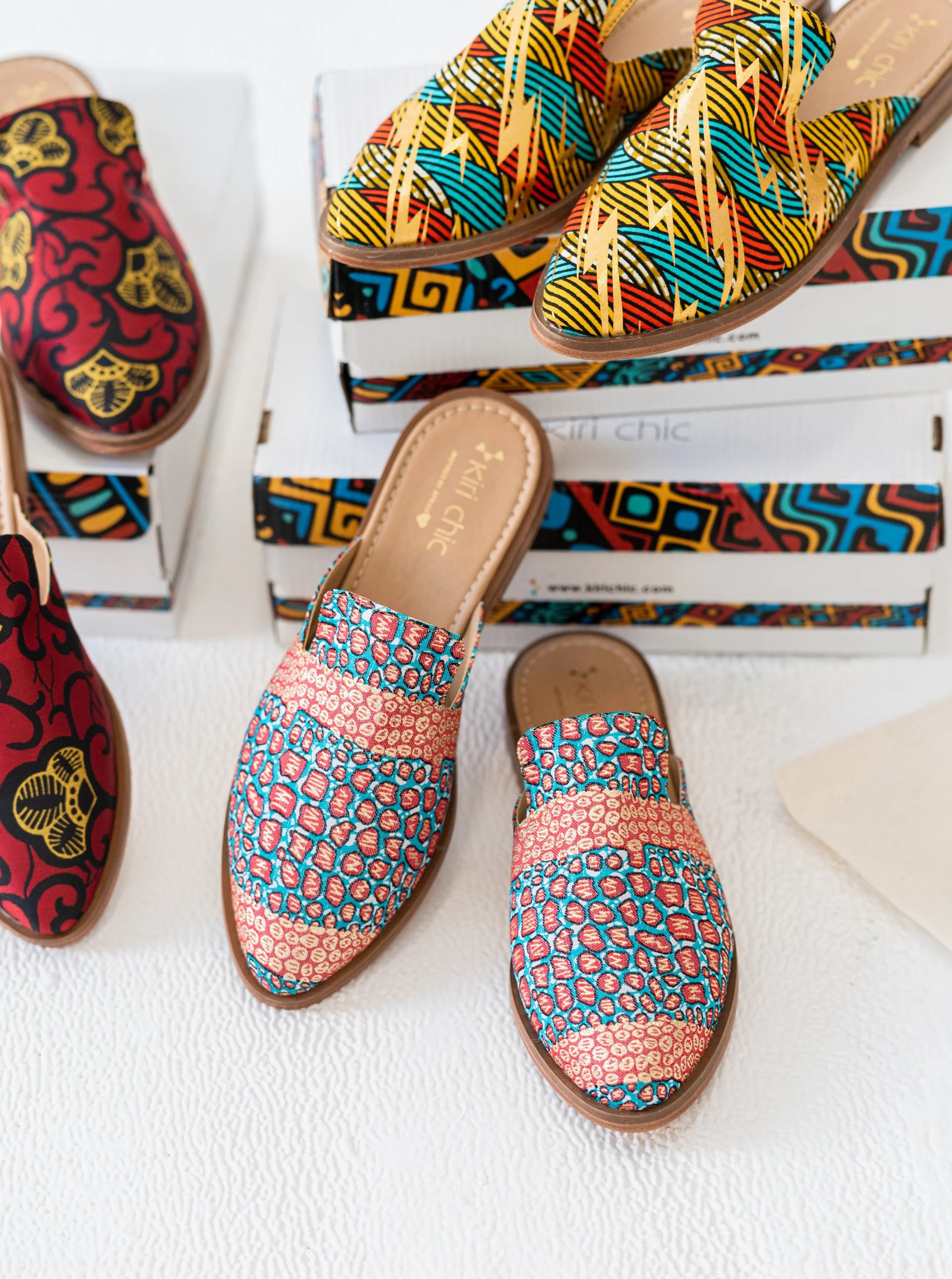 Colorful patterned shoes, including slip-on loafers with red, blue, and cream geometric designs and open-toe sandals with multicolored zigzag and lightning bolt patterns, displayed on a white surface with shoe boxes in the background.